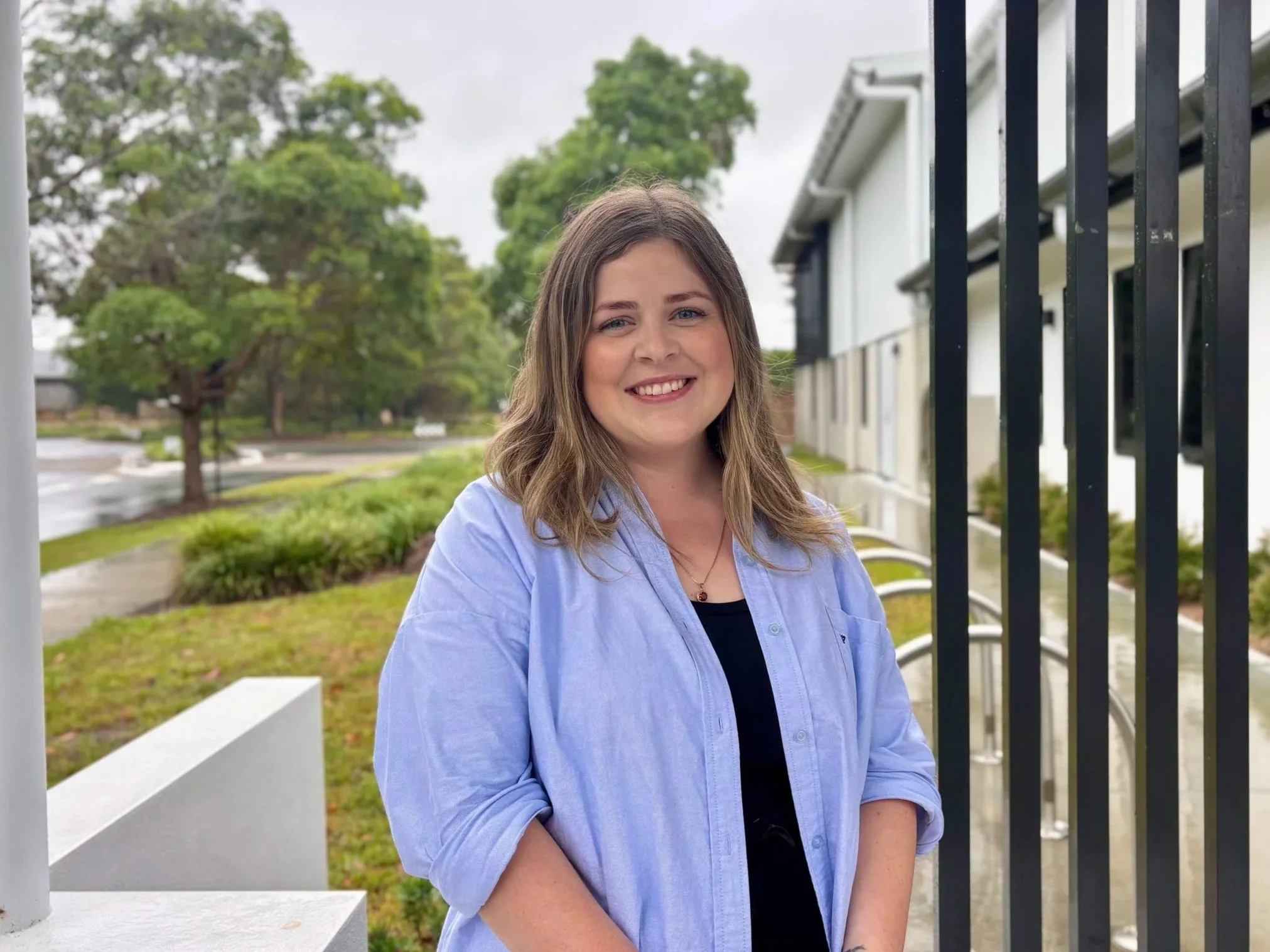 A young woman with shoulder-length brown hair, smiling, wearing a blue shirt over a black top, standing outside near a black metal fence with a residential building and trees in the background.