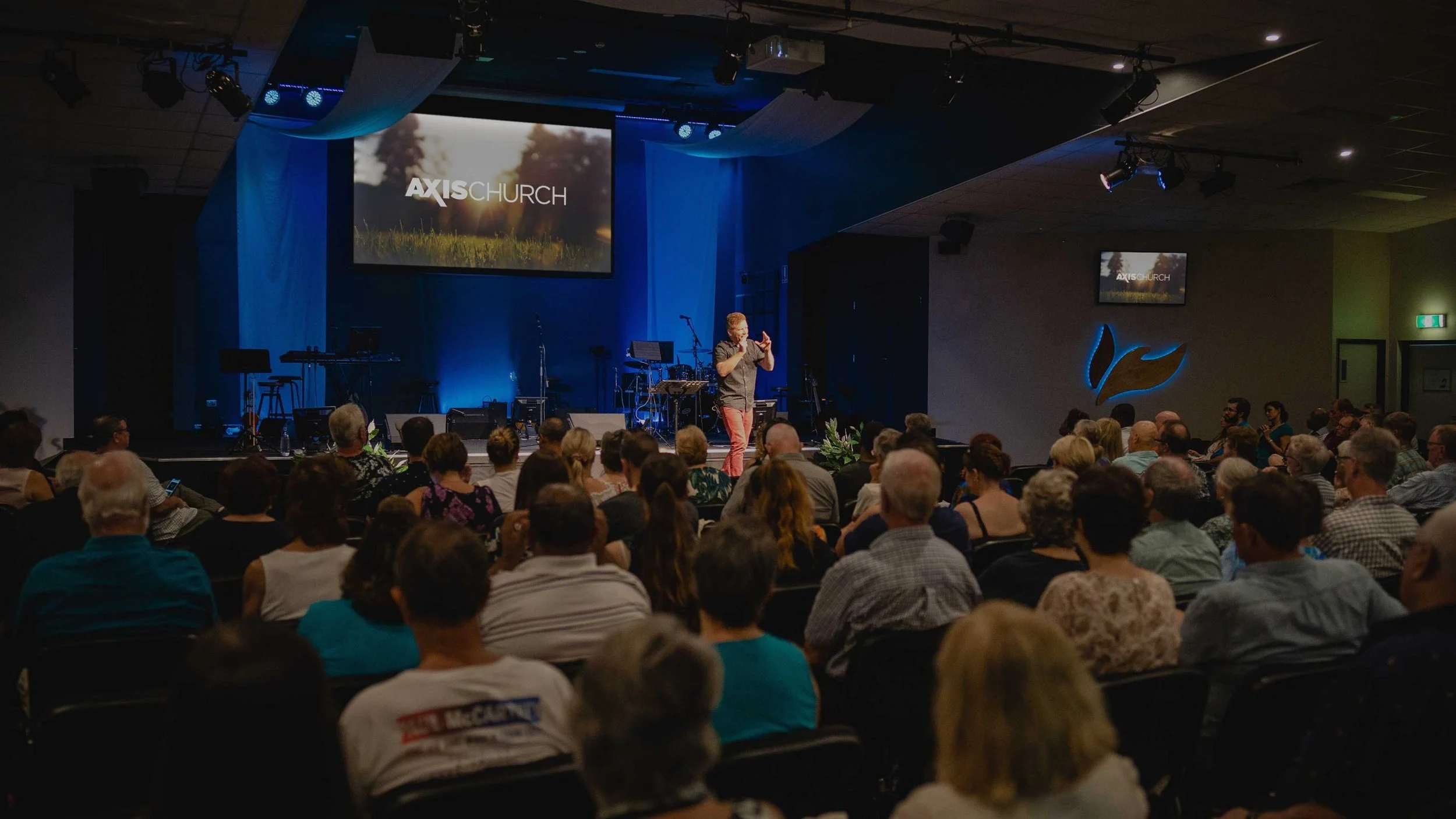 A speaker is presenting on stage at Axis Church to a large seated audience in a dark auditorium. The stage has musical instruments, and a large screen displaying 'AXISCHURCH' with a nature background. The audience includes a diverse group of people attentively listening.