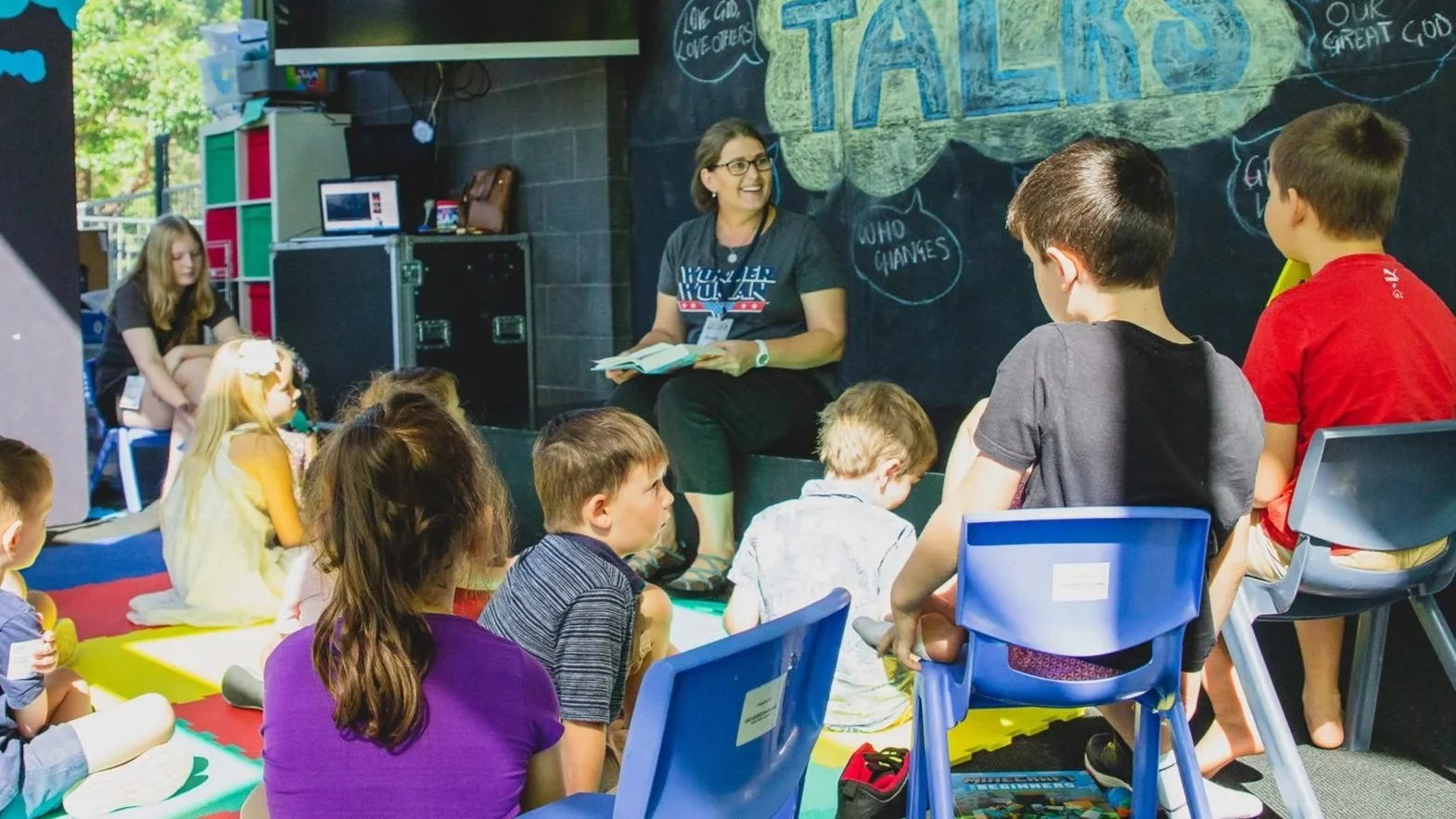 A group of children sitting on colorful mats and chairs in a classroom, listening to a woman teacher who is holding papers and smiling in front of a blackboard with chalk writing and drawings.