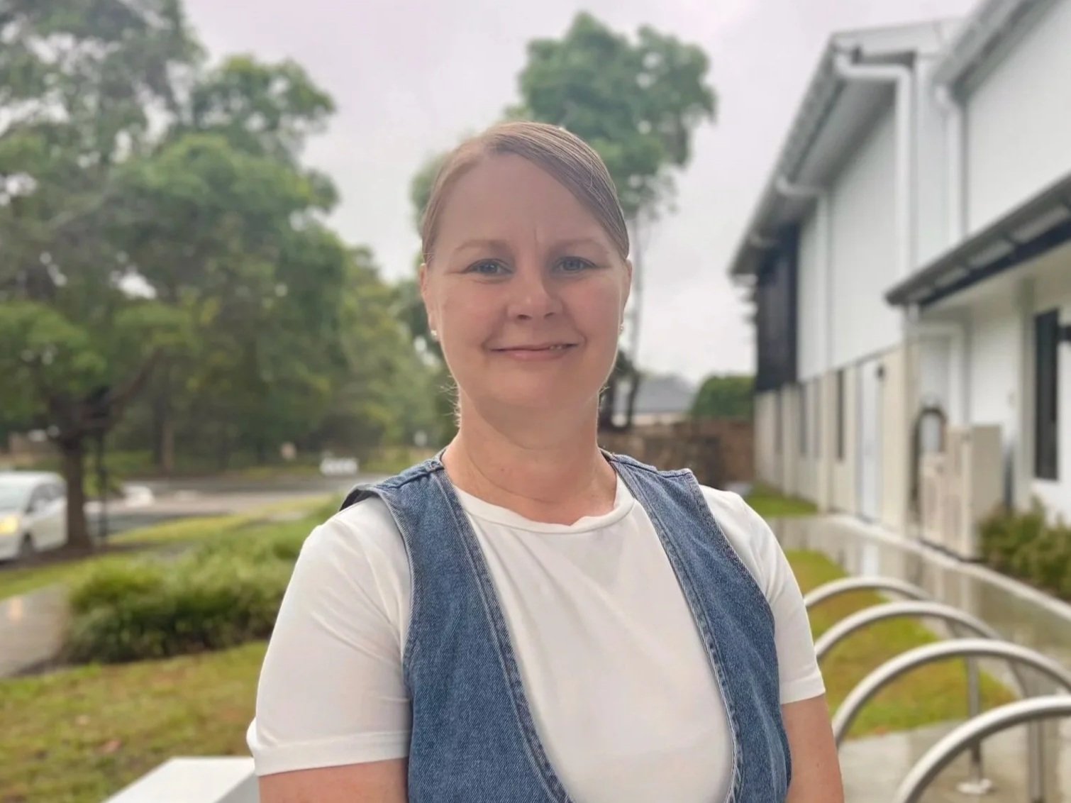 A woman with long, light brown hair, blue eyes, and fair skin, smiling and wearing a sleeveless dark blue top with a pattern, standing against a white brick wall with some green leaves in the upper right corner.