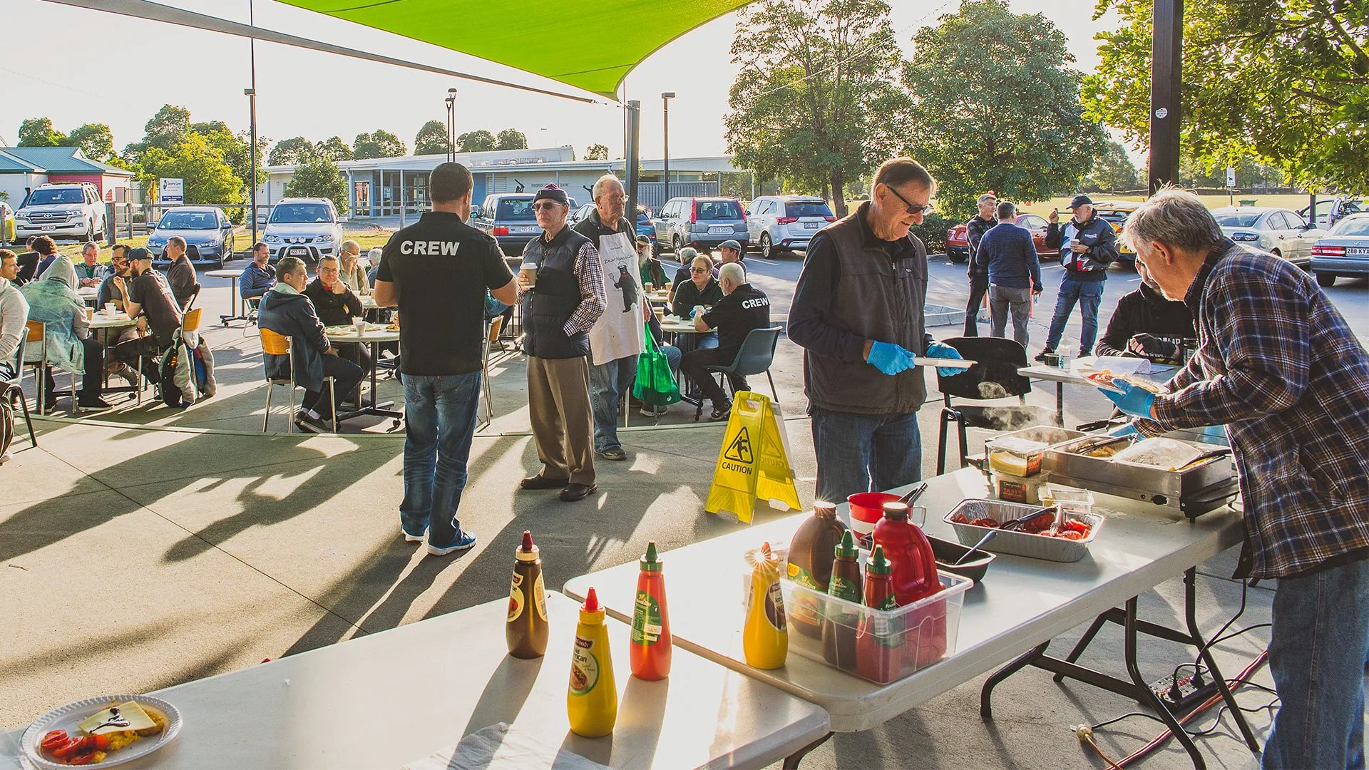 People gather at an outdoor event with a food service table, some people wearing shirts labeled 'CREW', and a man serving food from a chafing dish. There are tables with seating, a parking lot in the background, and a large patio umbrella providing shade.