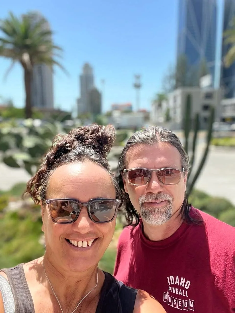 A smiling woman and a man wearing sunglasses taking a selfie outdoors with city buildings, palm trees, and cacti in the background on a sunny day.