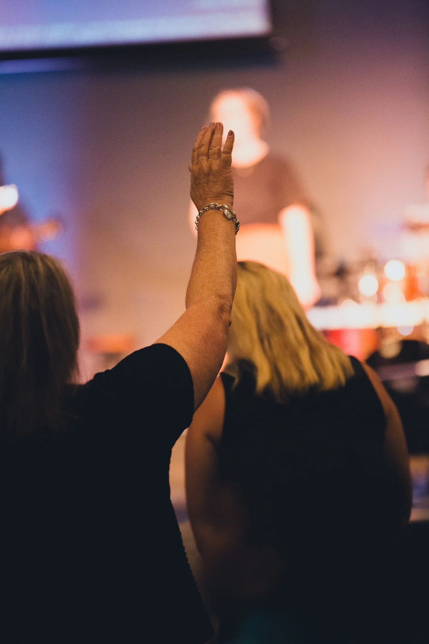 A woman with a bracelet on her wrist raises her hand during a concert or event, with a blurred stage and performer in the background.