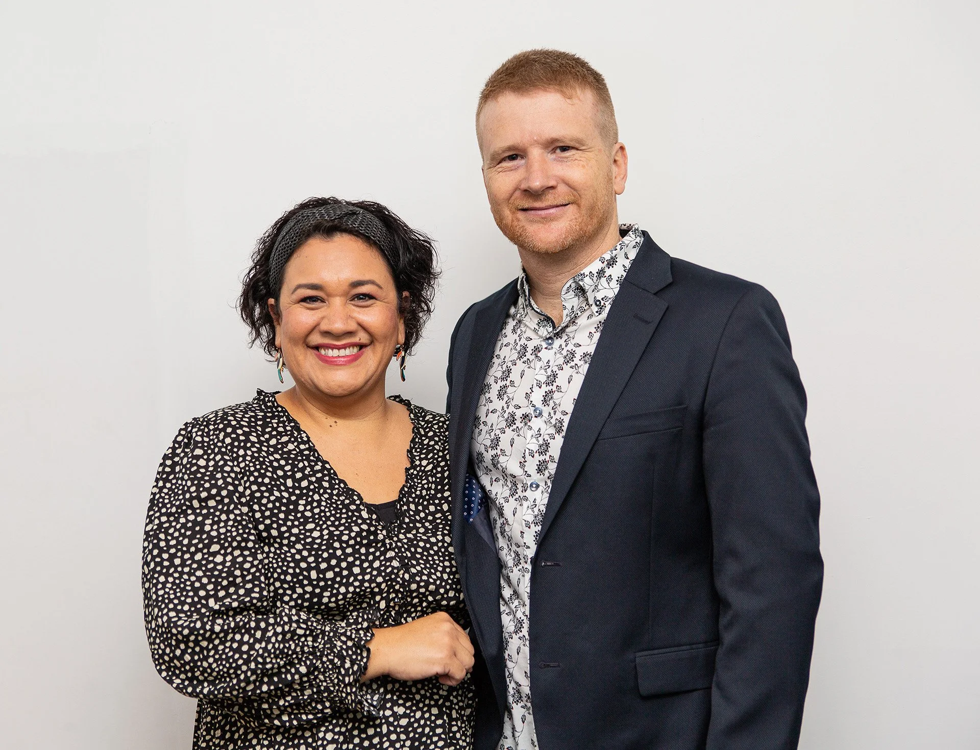 Two people standing together smiling in front of a plain white background, a woman on the left with curly dark hair, wearing a black and white patterned blouse, and a man on the right with short blonde hair and beard, wearing a dark blazer and a white shirt with a floral pattern.