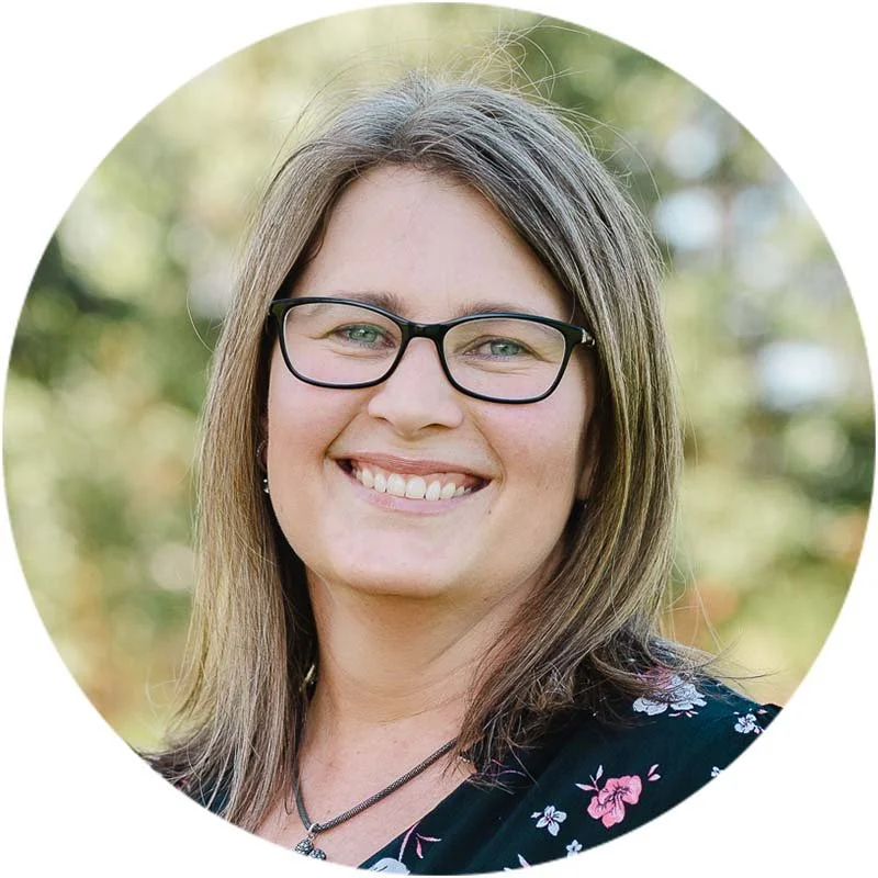 A woman with shoulder-length brown hair, glasses, and a floral top, smiling outdoors with a blurry green background.