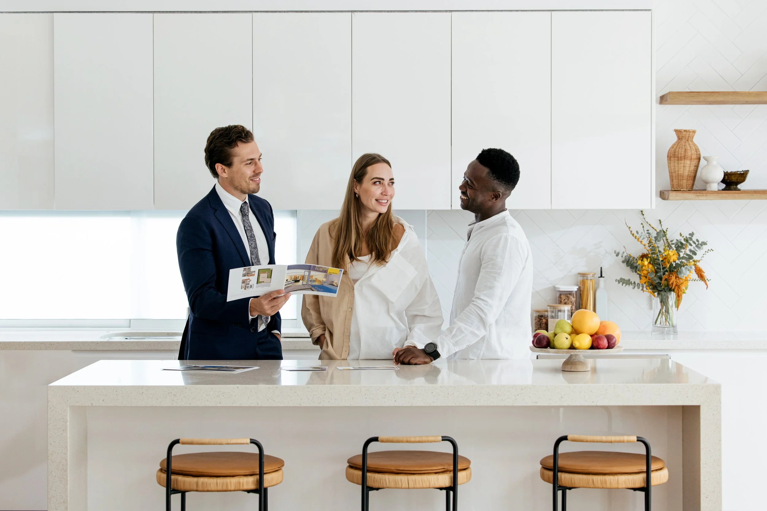 Agent in a suit holding a property magazine and consulting clients behind a kitchen island in a staged Bream Bay home. Sell your Bream Bay property with James Alexander and Charlotte Daun.