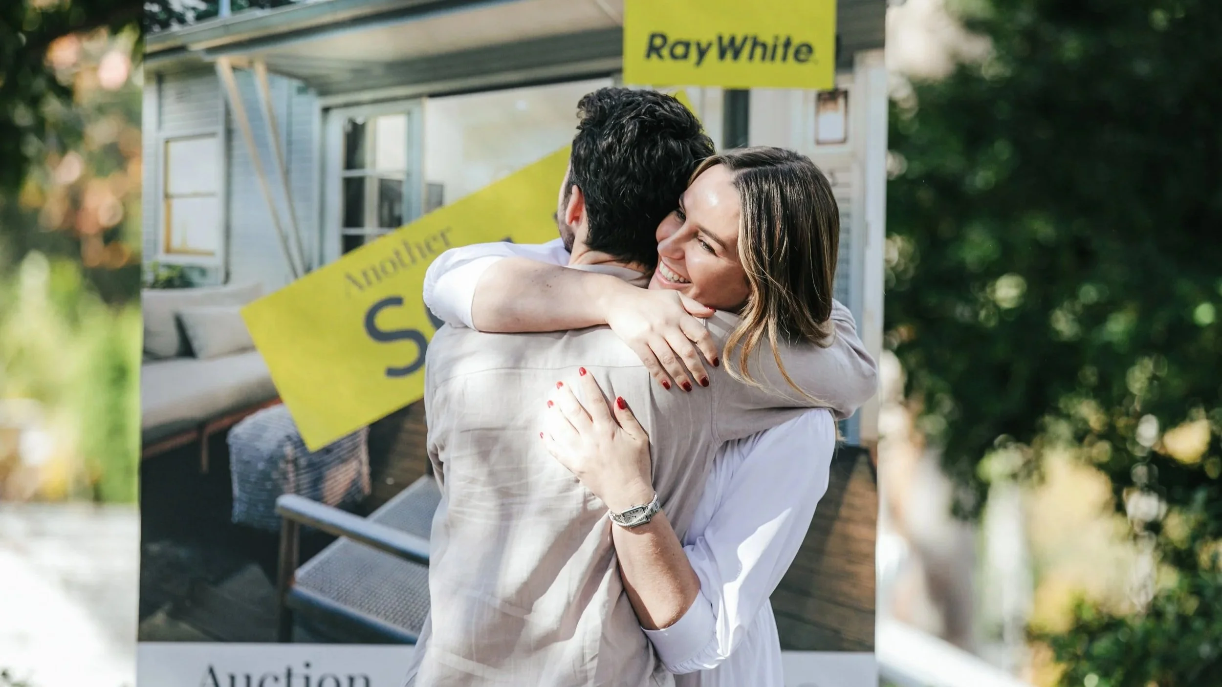 A smiling man and woman hugging in front of a house after a successful property auction. Bream Bay real estate experts James Alexander and Charlotte Daun.