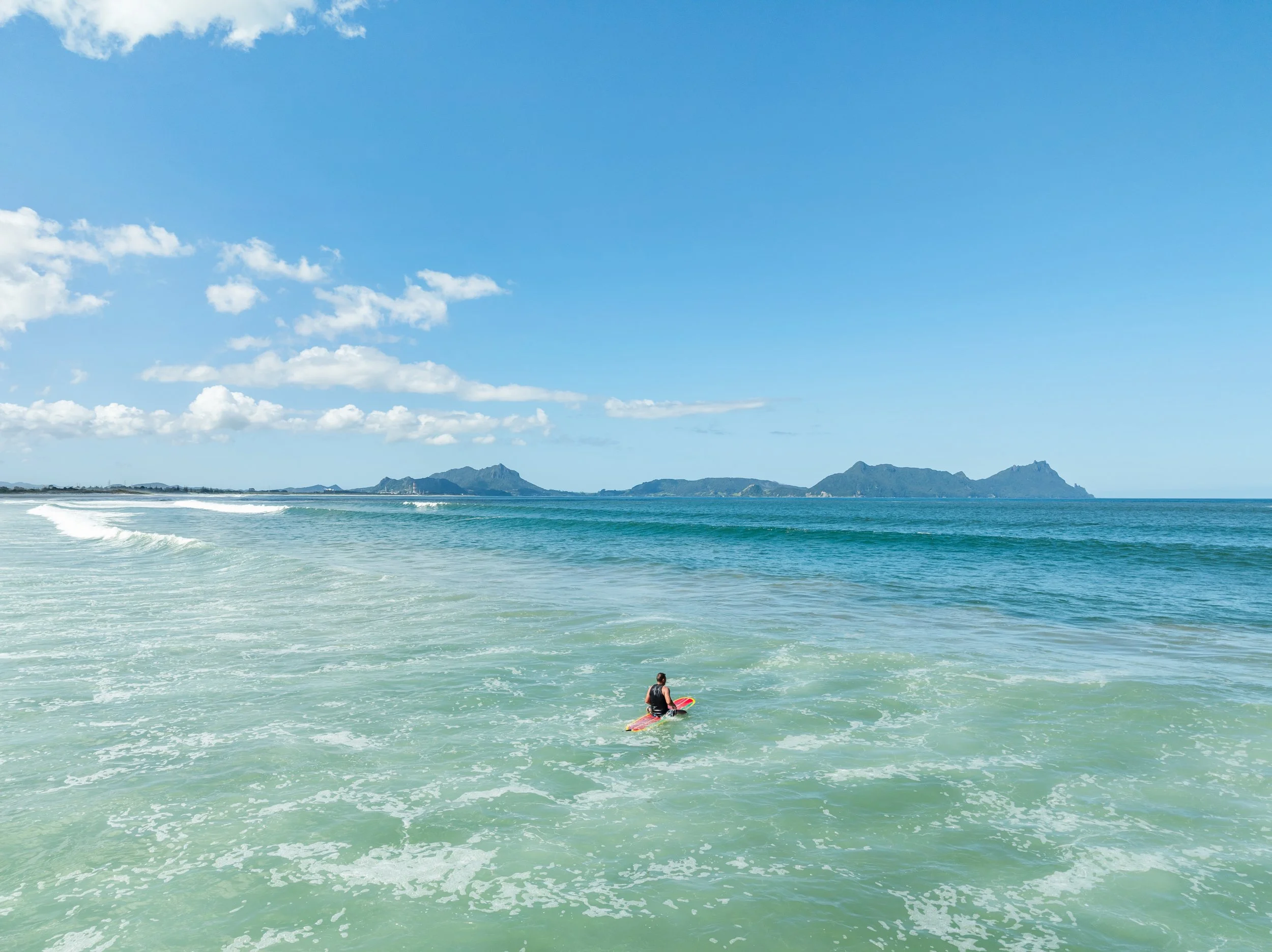 Sunny aerial view of a surfer in the waves at Ruakaka Beach looking towards Whangārei Heads, showcasing the active Bream Bay lifestyle. Sell your Bream Bay property with James Alexander and Charlotte Daun.