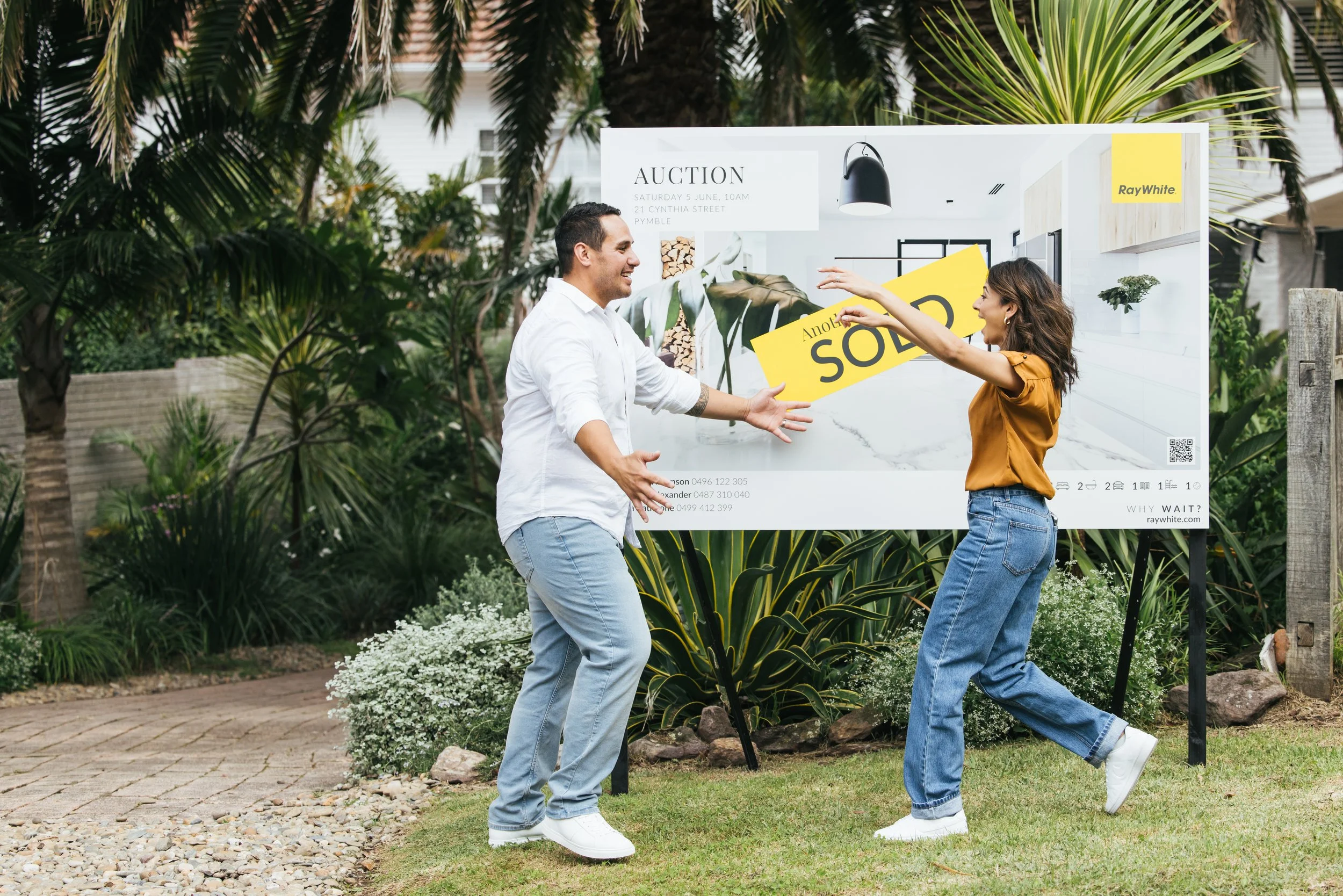 Happy clients celebrating a successful property sale next to a yellow Ray White SOLD signboard. Sell your Bream Bay property with James Alexander and Charlotte Daun.