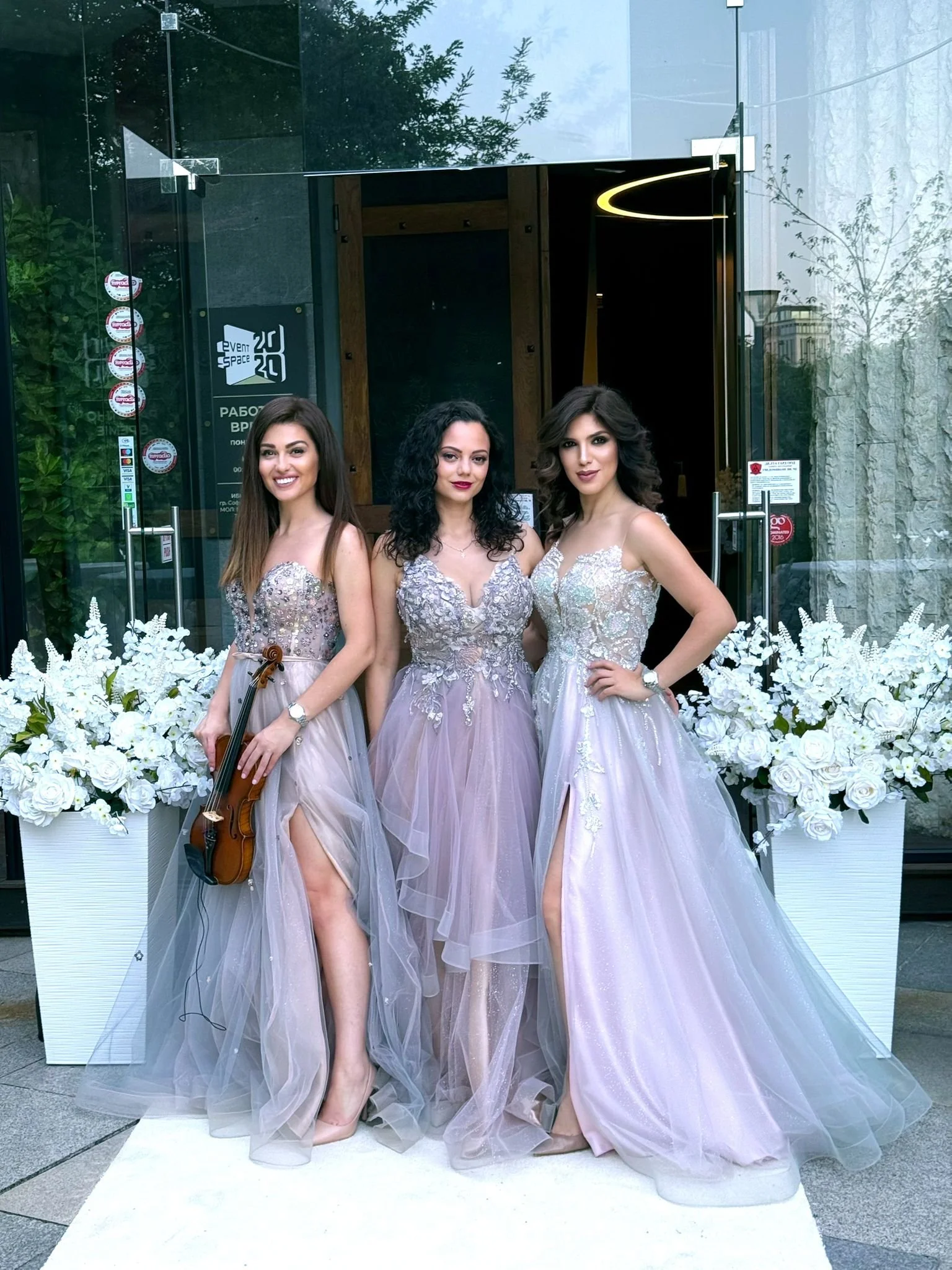 Three women in elegant lavender and silver evening gowns standing in front of a floral display and glass entrance, one holding a violin.