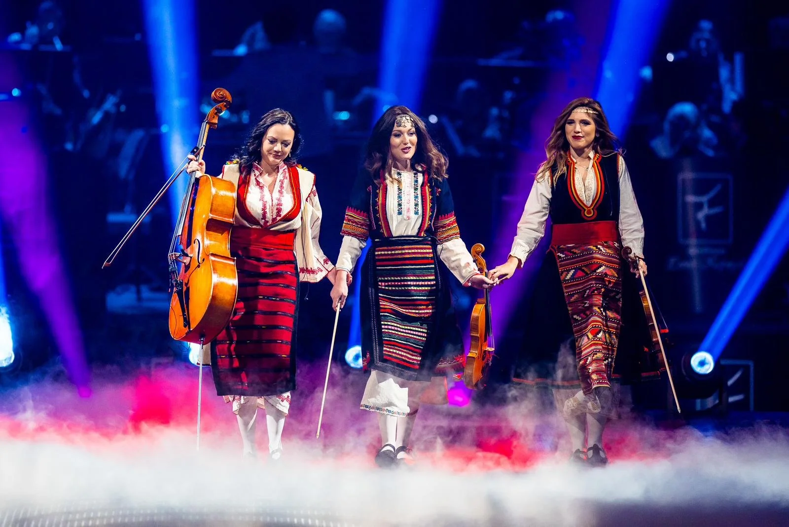 Three women in traditional Eastern European folk costumes on stage, holding musical instruments, under vibrant stage lights with smoke effects.