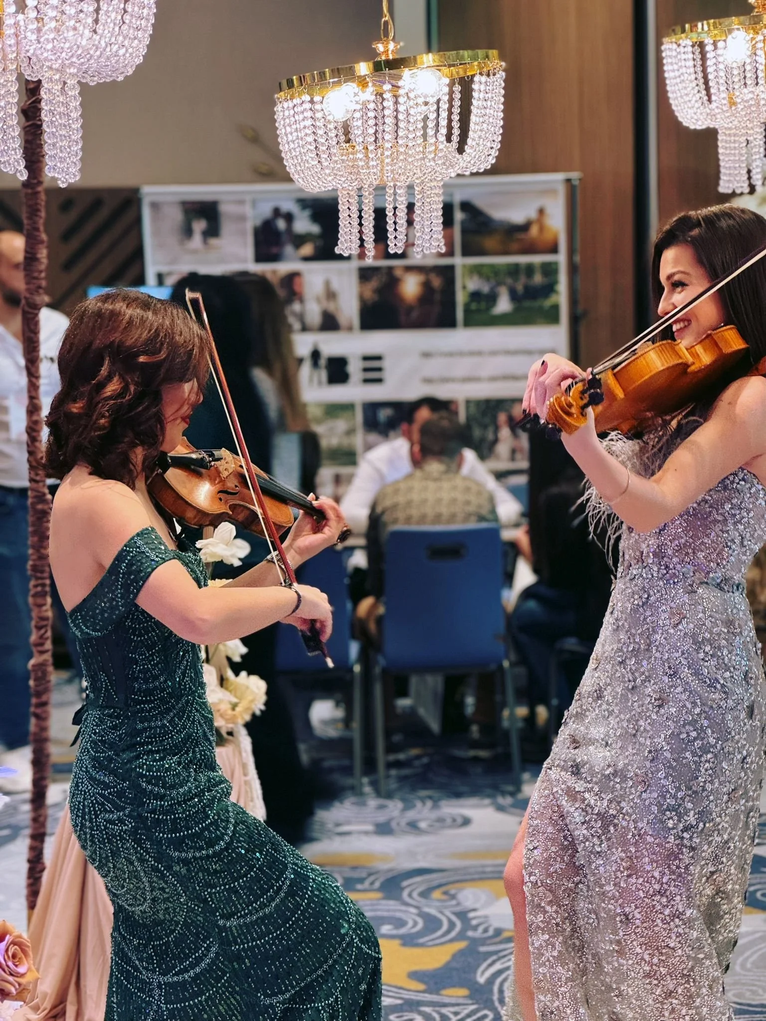 Two women in elegant dresses playing violins at an indoor event with chandeliers and a decorated background.