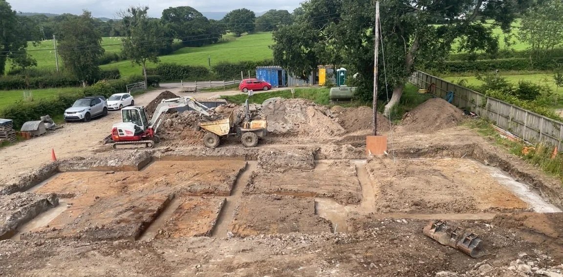 Construction site with excavated foundation trenches, small bulldozer, a mini-excavator, and parked cars in the background.