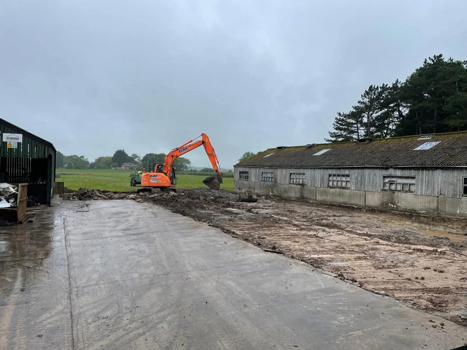 An orange excavator is working next to a weathered gray barn on a cloudy, rainy day, with a dirt area being prepared or excavated.