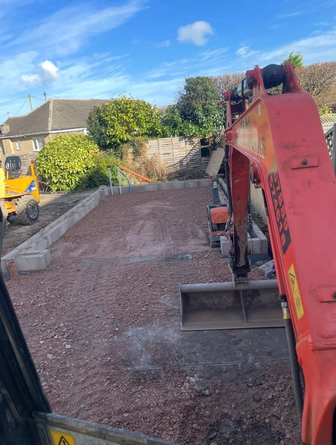 A construction site with a small excavator on a gravel surface, red and yellow machinery, and a residential area in the background with trees and a wooden fence under a partly cloudy sky.