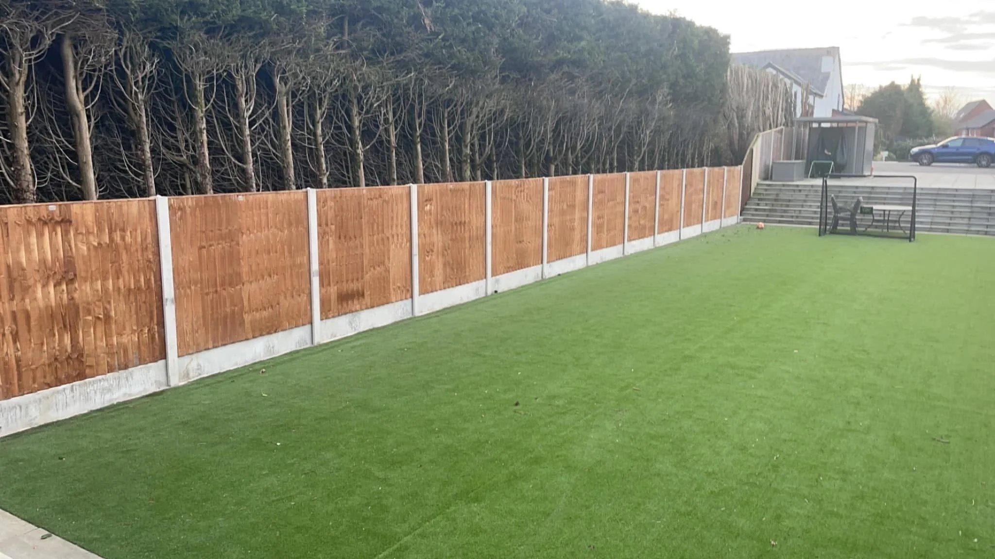 A backyard with a green artificial lawn, a wooden fence, and a staircase leading to an outdoor seating area with black chairs. There is a small gate at the back and some trees along the fence. The sky is partly cloudy.