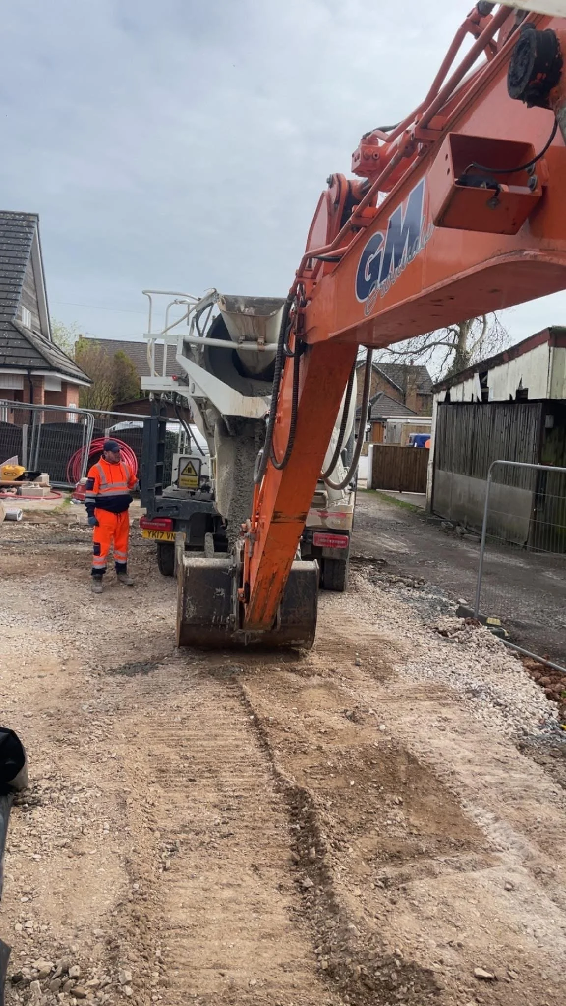 Construction site with a large orange excavator moving dirt and gravel. A worker in orange safety gear and blue jacket stands nearby, observing the work.