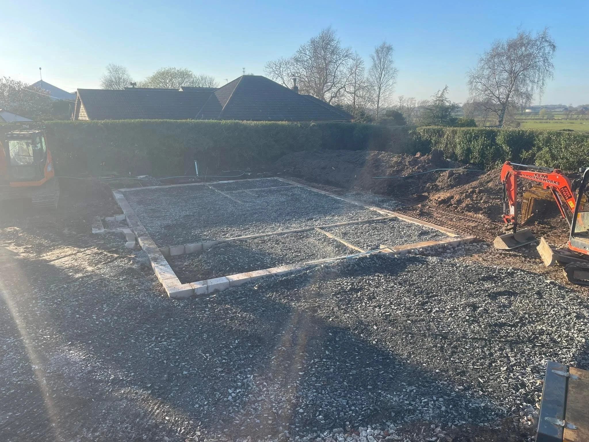 Construction site with foundation work in progress, gravel base laid out within boundary marked by concrete blocks, small excavator on the right.