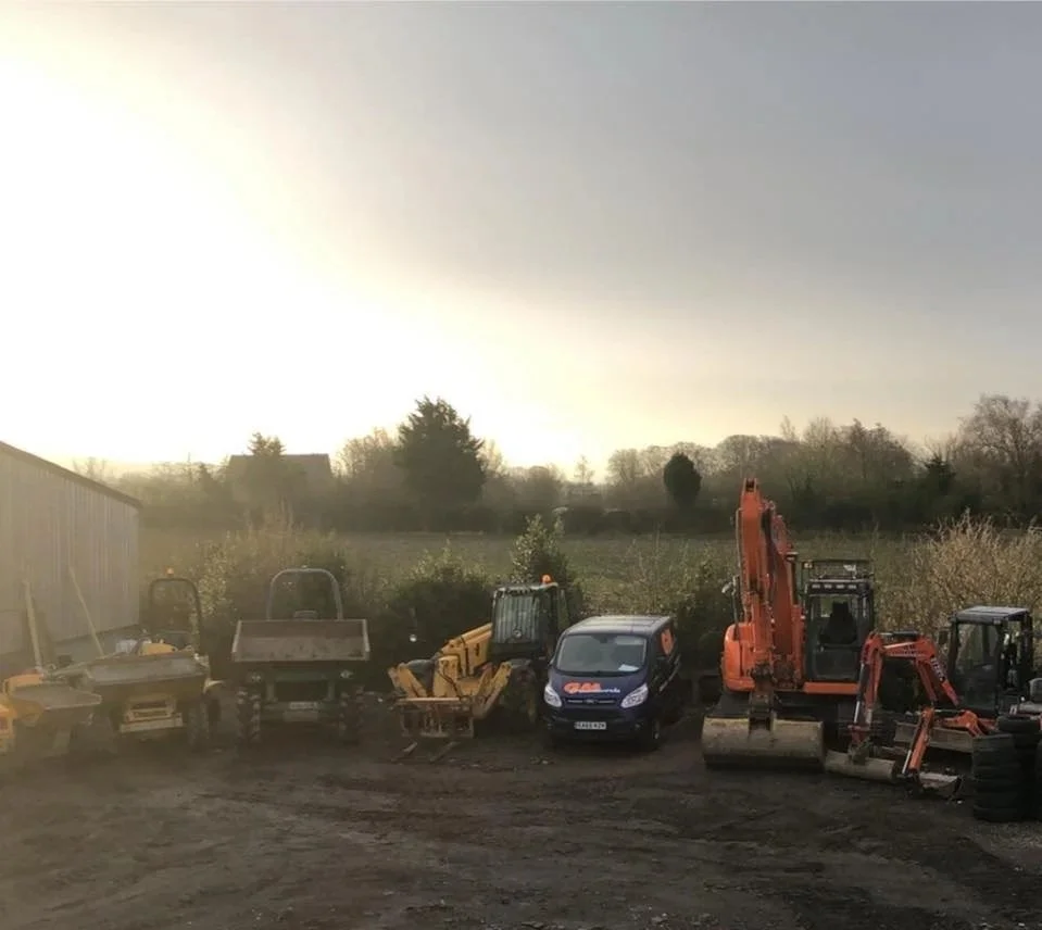 Construction equipment and vehicles, including excavators, and a van, lined up on a dirt lot with trees and a cloudy sky in the background.