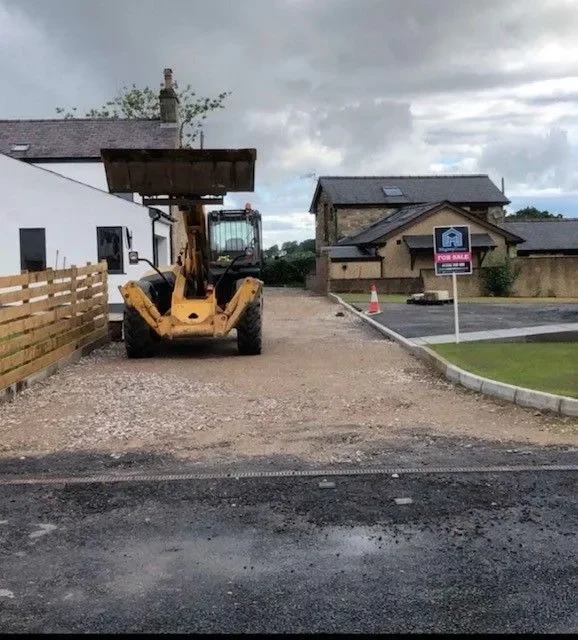 A construction vehicle, possibly a forklift, parked on a gravel driveway in front of a white building with a wooden fence on the left. There are residential houses and a parking lot with a 'For Sale' sign on the right. Overcast sky.