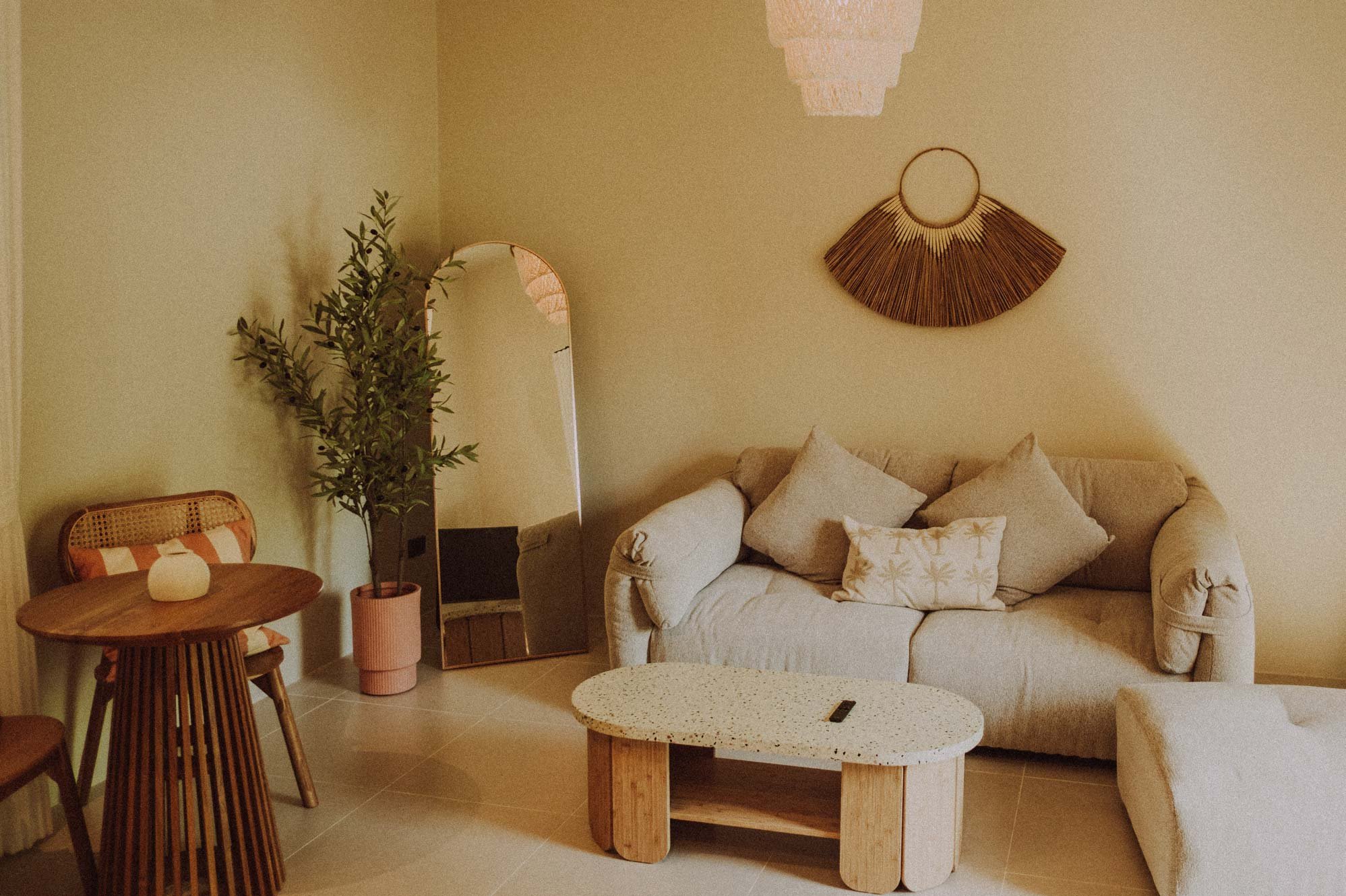 A cozy living room with beige walls and furnishings, including a cream-colored sofa with pillows, a small wooden side table, a standing mirror, a tall plant in a pink pot, and wall decor made of natural materials.