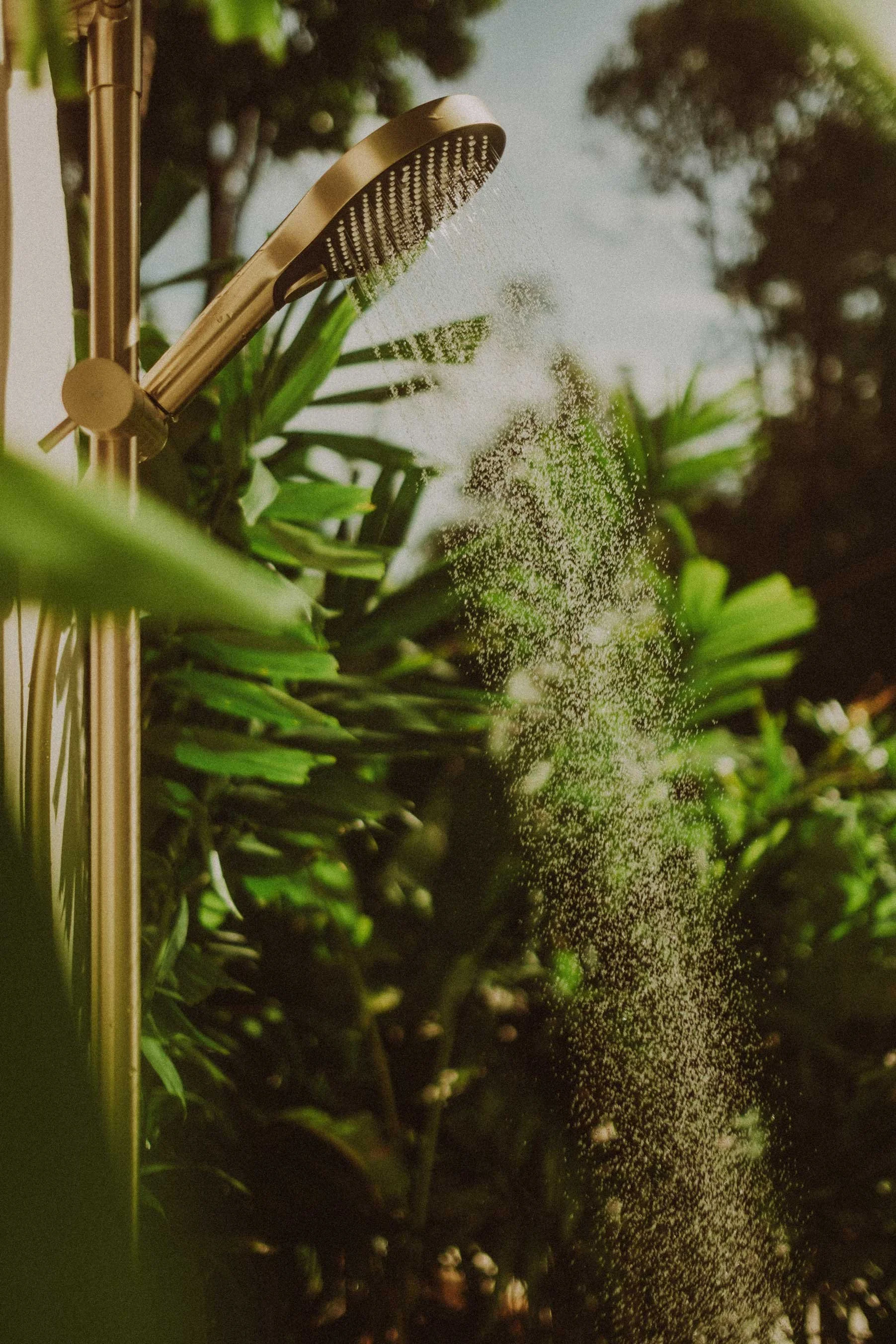 An outdoor shower with a gold-colored showerhead spraying water, surrounded by lush green tropical plants and trees in the background.