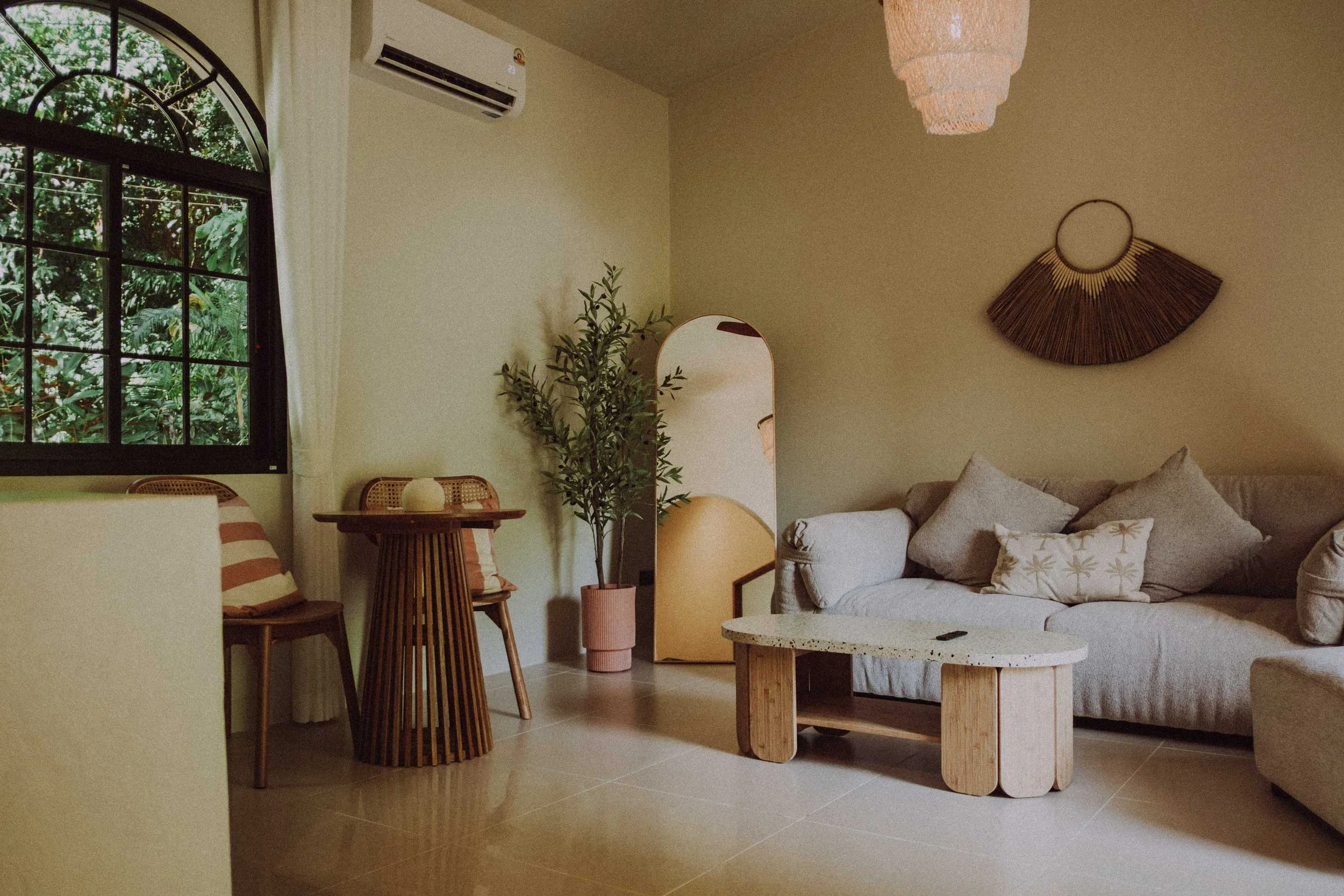 Living room with beige sofa, lattice side table, wooden chairs, small table, potted plant, mirror, window with trees outside, air conditioner, pendant light, and wall art.