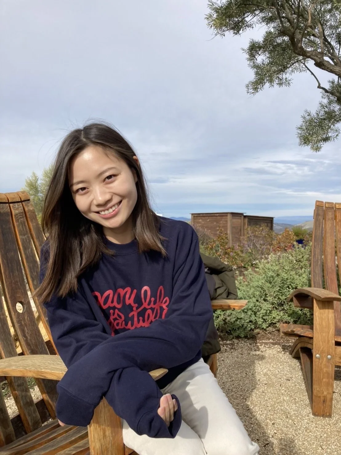 A young woman with long brown hair smiling, sitting outdoors on wooden benches, with a cloudy sky, greenery, and distant hills in the background.