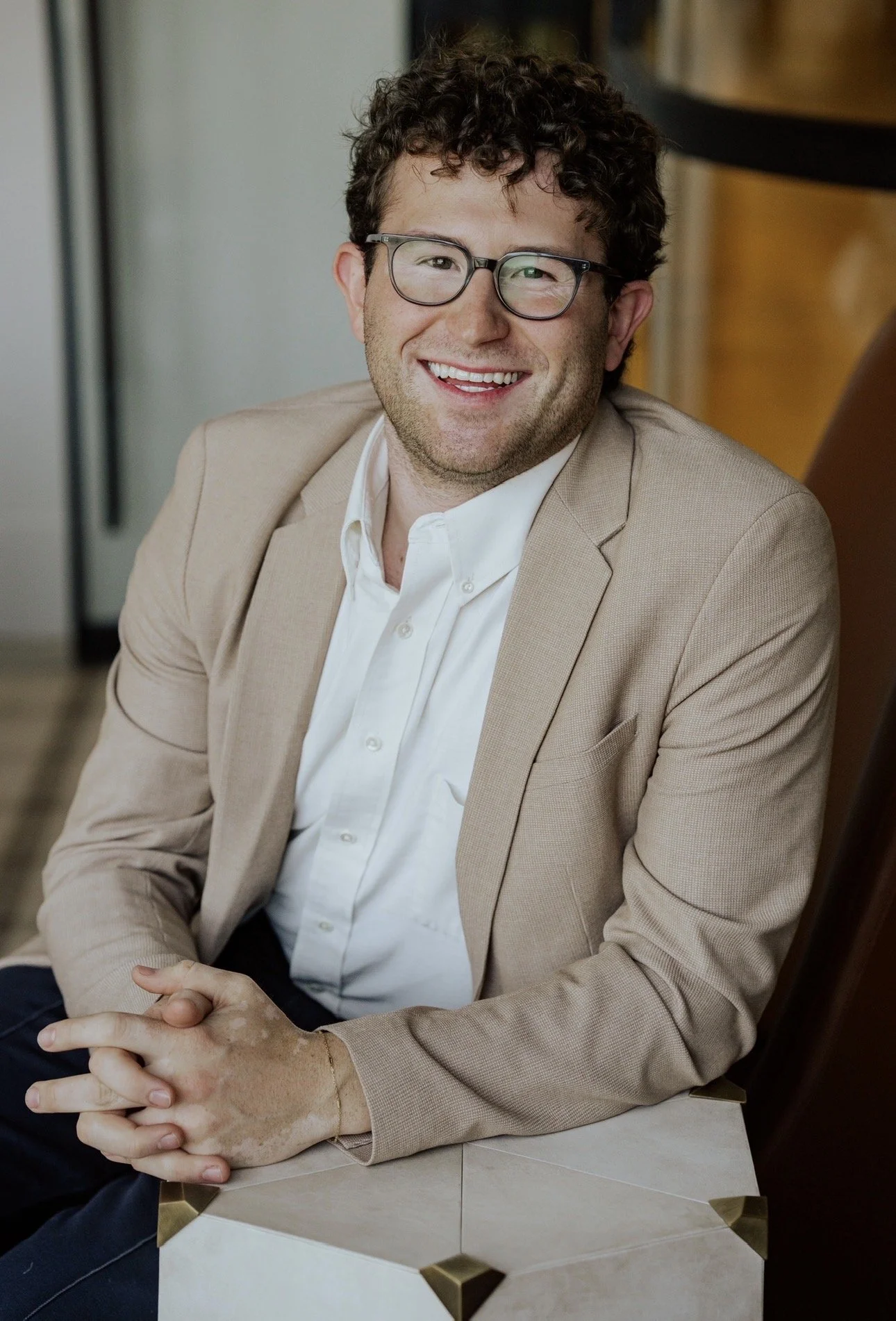 Smiling man with curly hair, glasses, wearing a beige blazer and a white shirt, sitting at a table with hands clasped, in a professional setting.