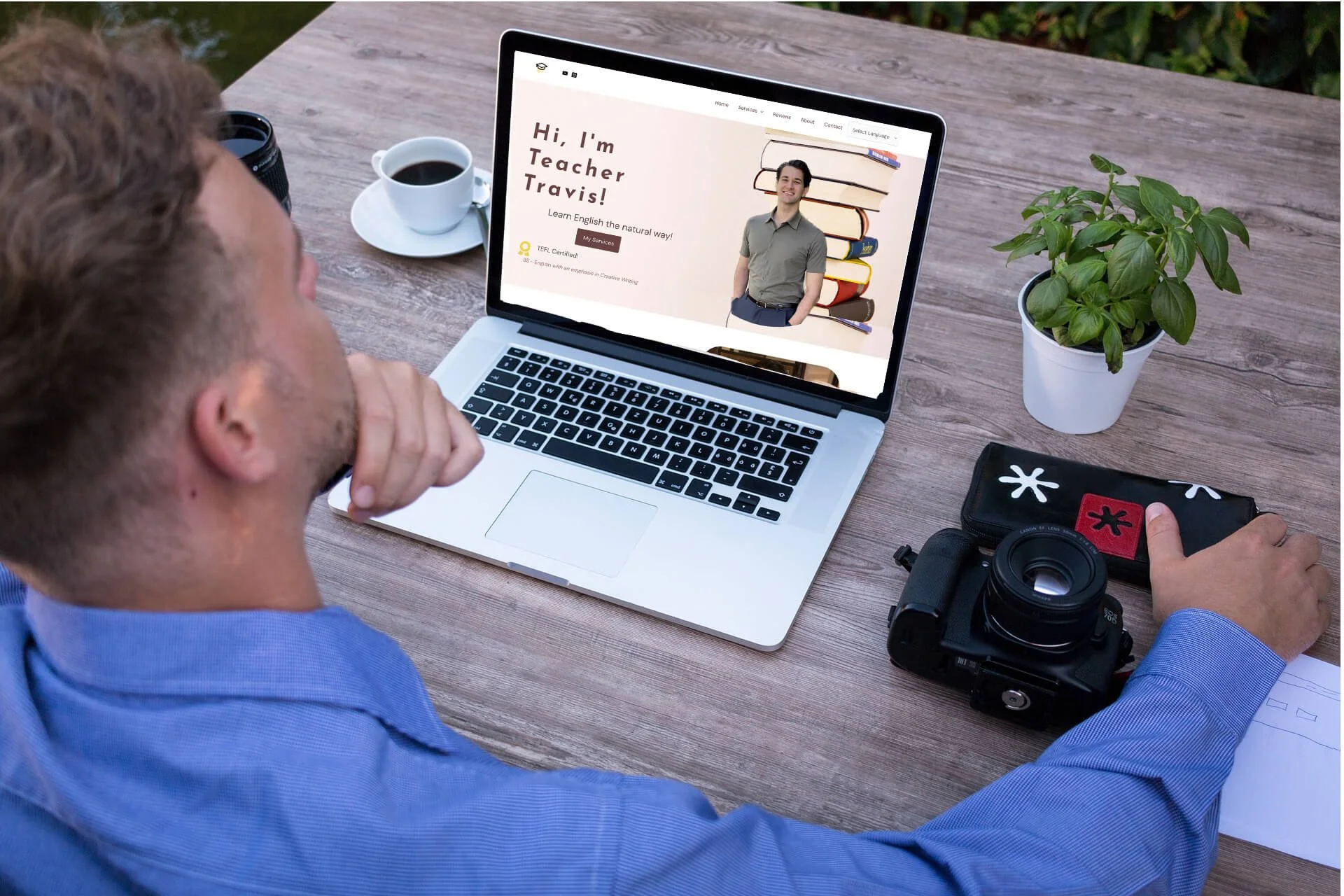 A man in a blue shirt sitting at a wooden table outdoors looking at a laptop screen displaying a website for an English teacher named Travi, with a profile photo of him, a stack of books, and text about learning English naturally.