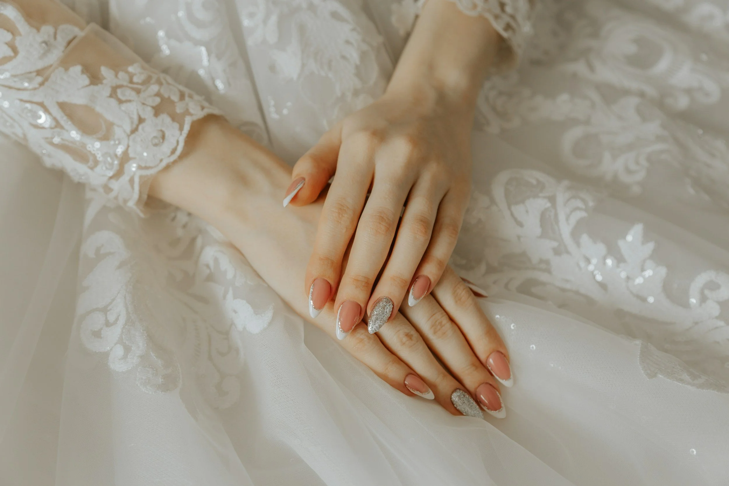 Close-up of a bride's hands with manicured nails resting on white lace fabric of her wedding dress.