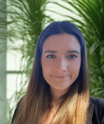 A psychologist with long brown hair smiling in front of green leafy plants.