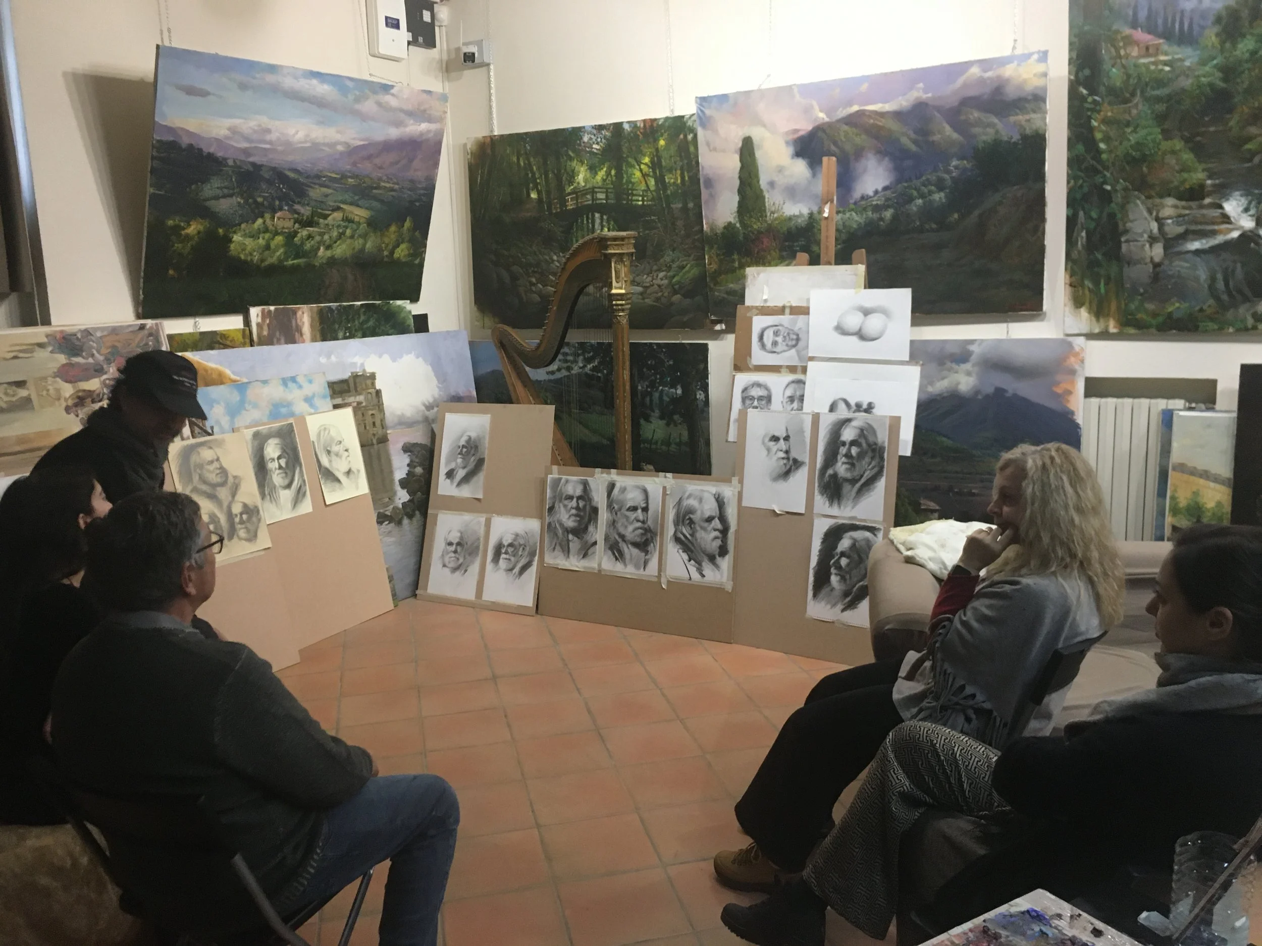 Una stanza con molte opere d'arte, tra cui televisioni, quadri e disegni di volti e paesaggi. Un gruppo di persone si siede e osserva gli artisti mentre mostrano i loro lavori.