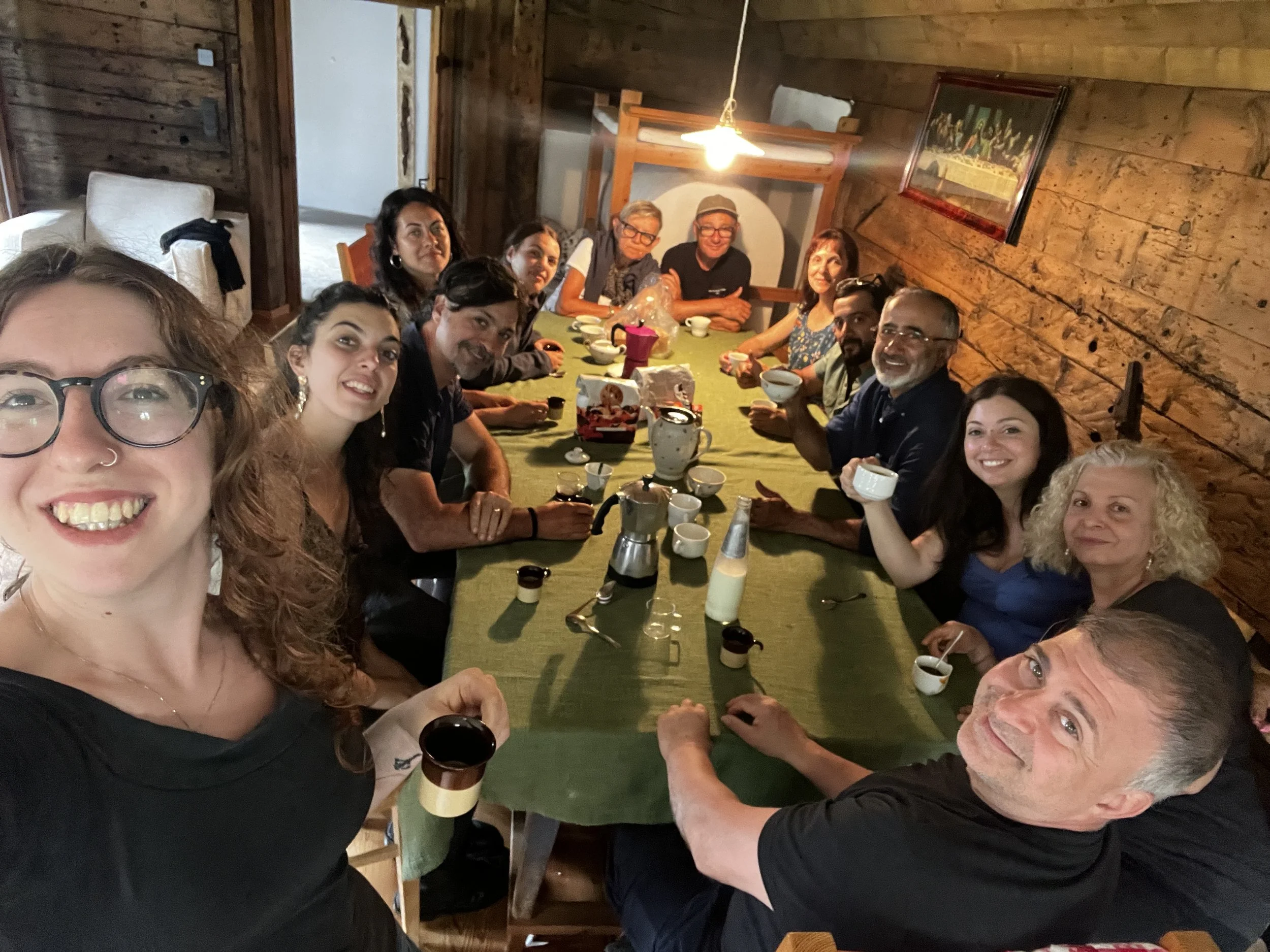 Un gruppo di persone sedute intorno a un tavolo in una stanza di legno, che brindano con tazze e ciotole, sorridendo alla fotocamera.