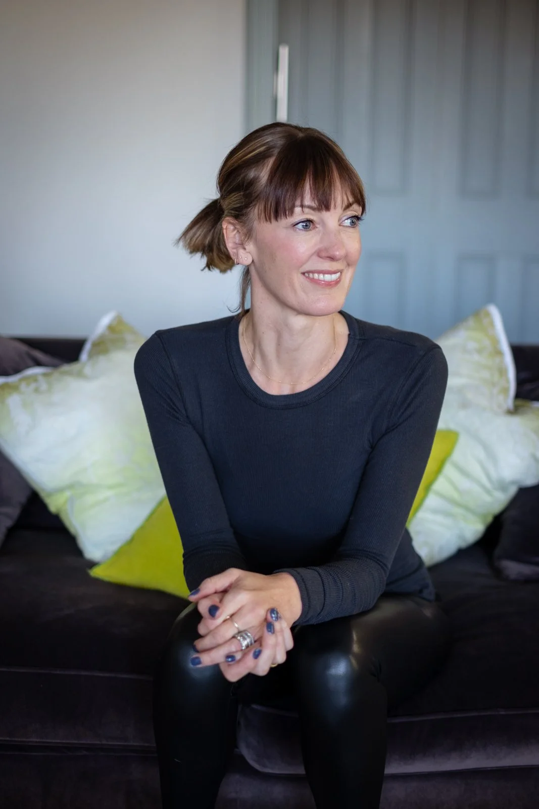 A woman with short brown hair and bangs sitting on a dark sofa with white and lime green pillows, smiling and looking to her right.
