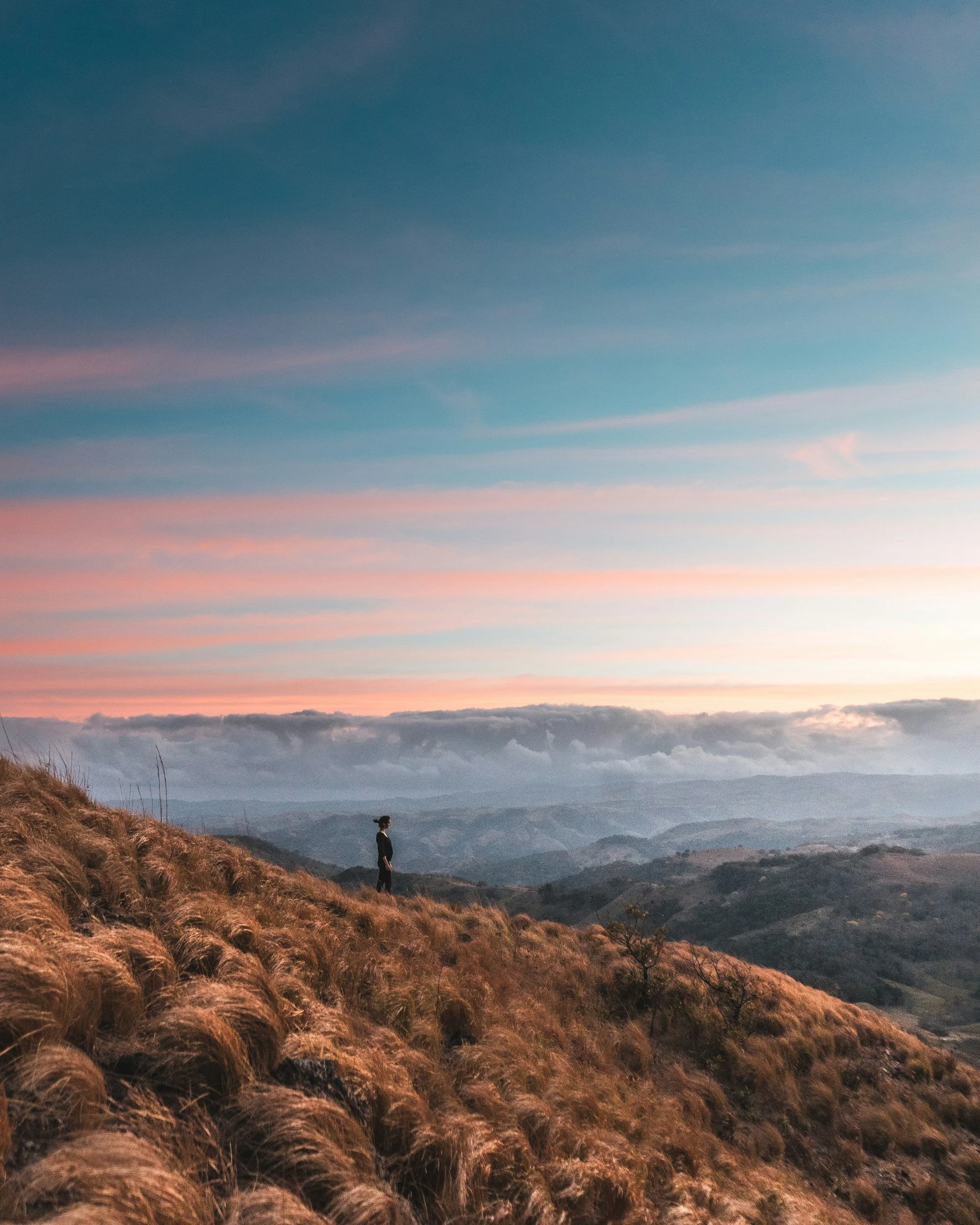 A person standing on a hillside covered with dry grass, overlooking a mountain landscape at sunset with a colorful sky and scattered clouds.