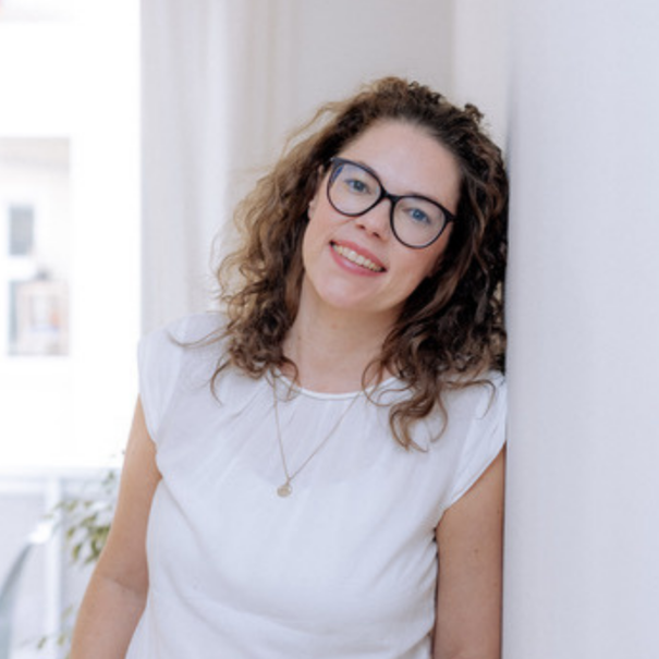 Lächelnde junge Frau mit Brille und lockigem Haar, trägt weißes T-Shirt, steht an einer weißen Wand in hell erleuchteter Umgebung.