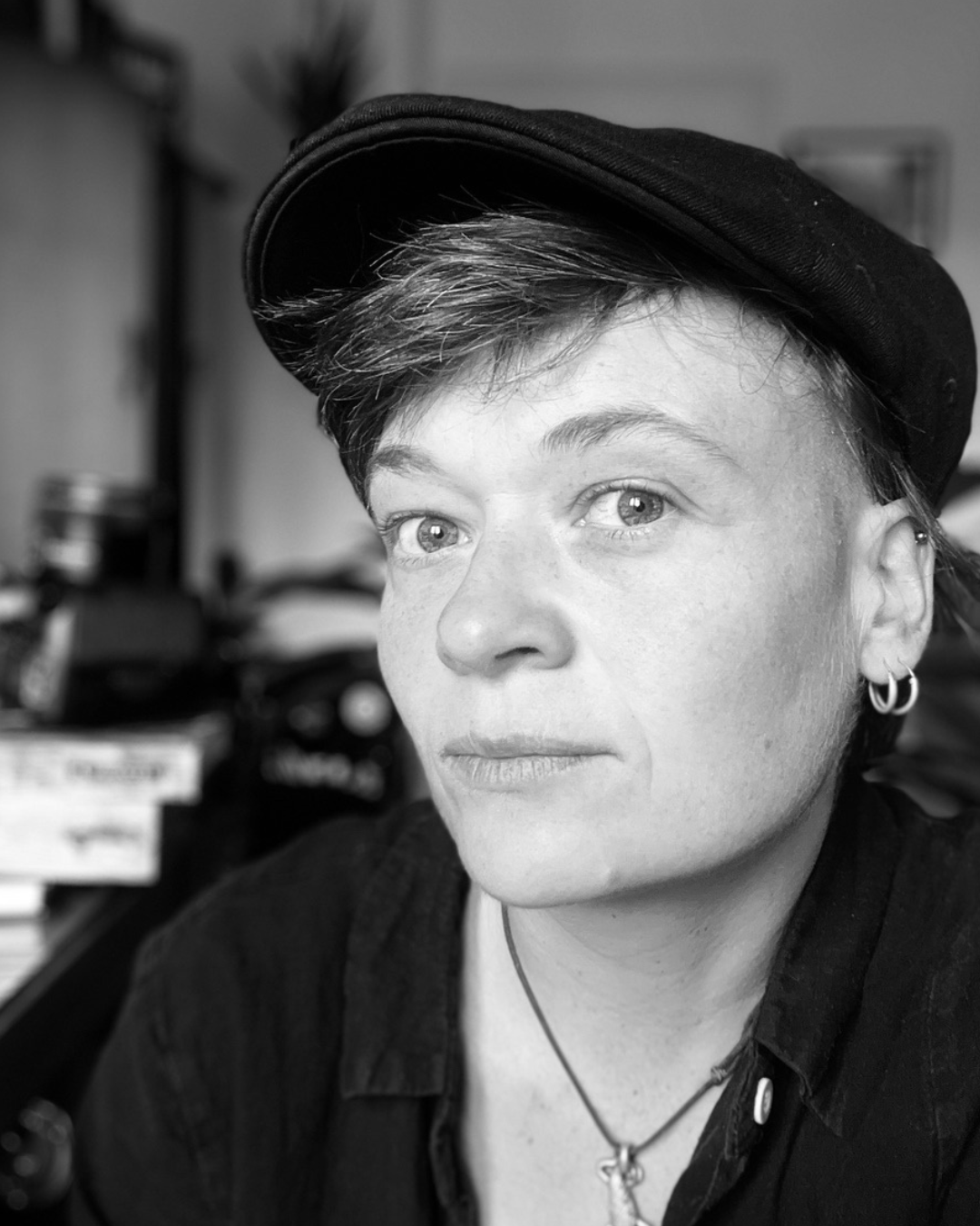 Schwarz-Weiß-Porträt einer jungen Frau mit kurzen Haaren, die eine schwarze Kappe trägt, mehrere Piercings, Make-up und Ohrringe trägt, im Hintergrund sind Bücher und Gegenstände sichtbar.
