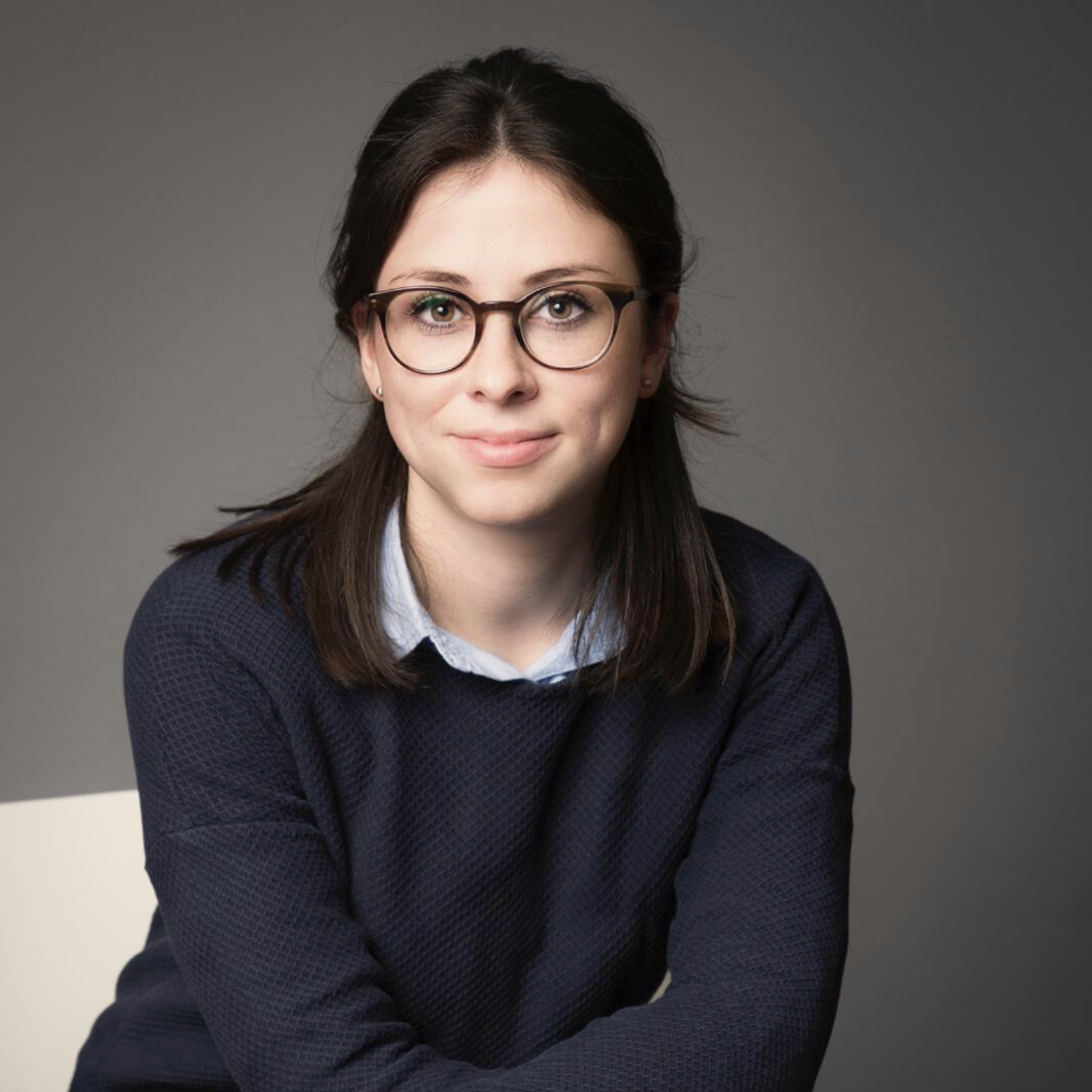 Porträt einer jungen Frau mit dunklem Haar, Brille, hellblauem Hemd und dunklem Pullover vor grauem Hintergrund.