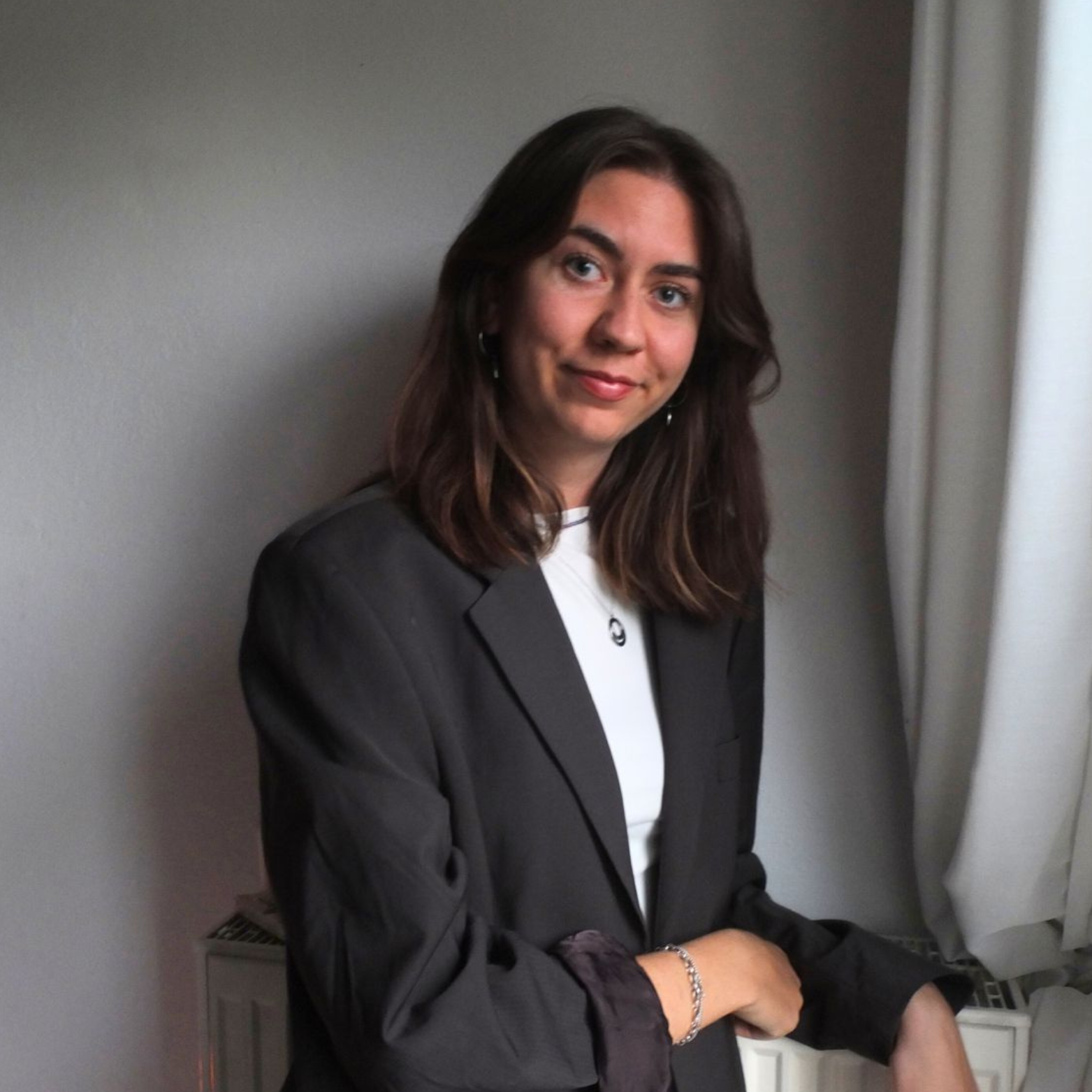 Junge Frau mit braunen Haaren, dunklem Anzug und weißen Shirt steht vor einem grauen Hintergrund neben einem Fenster mit weißen Vorhängen.