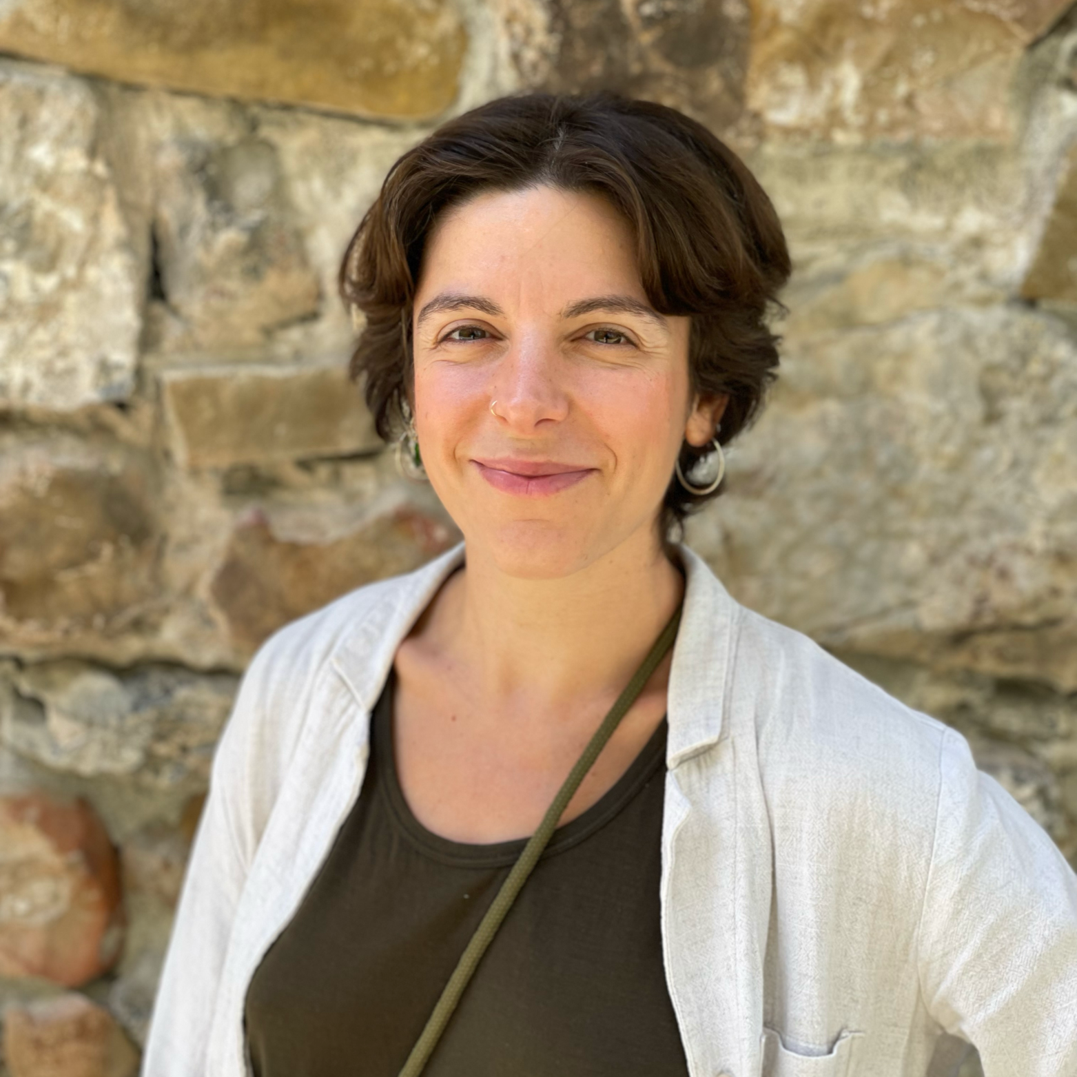 Eine Frau mit kurzen braunen Haaren, die vor einer Steinmauer steht, lächelnd in die Kamera schaut.