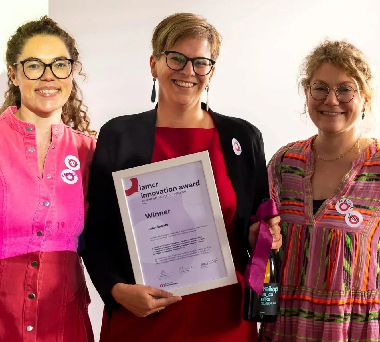 Drei Frauen stehen zusammen, eine hält ein Zertifikat für den iamcr Innovation Award, die mittlere Frau trägt eine schwarze Jacke und rote Kleidung, die rechte Frau trägt ein bunt gestreiftes Kleid, alle lachen und tragen Buttons.