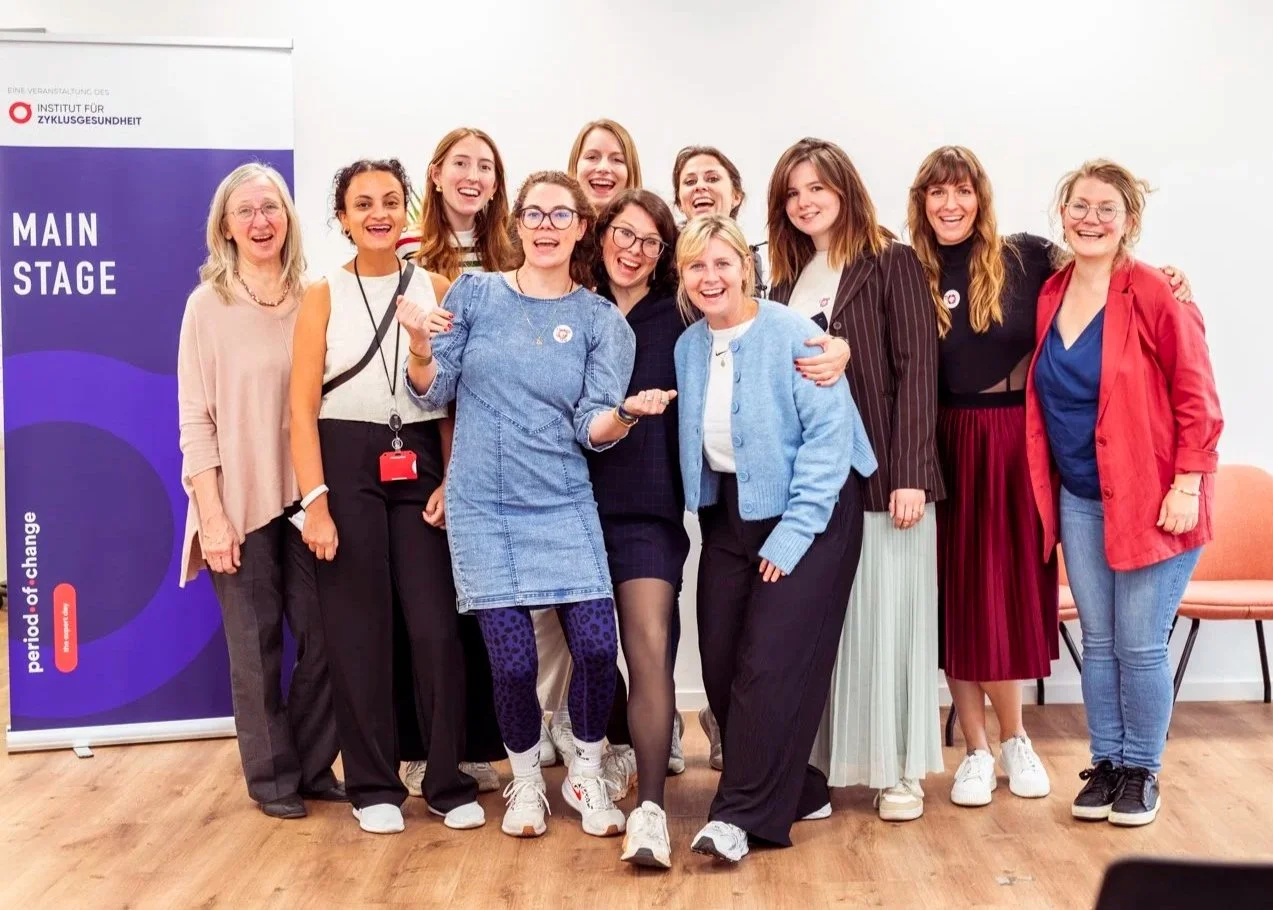 Gruppe von Frauen, die bei einer Veranstaltung des Instituts für Zyklusgesundheit vor einem Banner stehen, alle lachen und posieren zusammen.