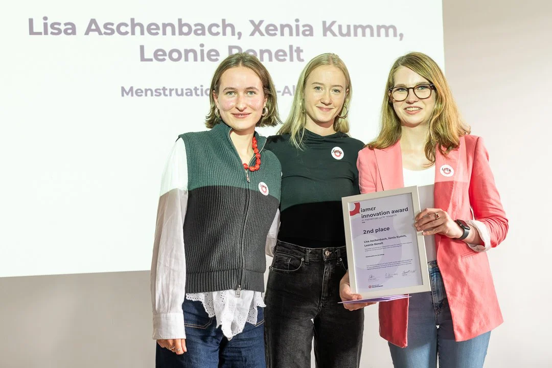 Drei Frauen stehen bei einer Preisverleihung, eine hält eine Urkunde, im Hintergrund ein Bildschirm mit Text, der die Namen Lisa Aschenbach, Xenia Kumm und Leonie Penelt zeigt.