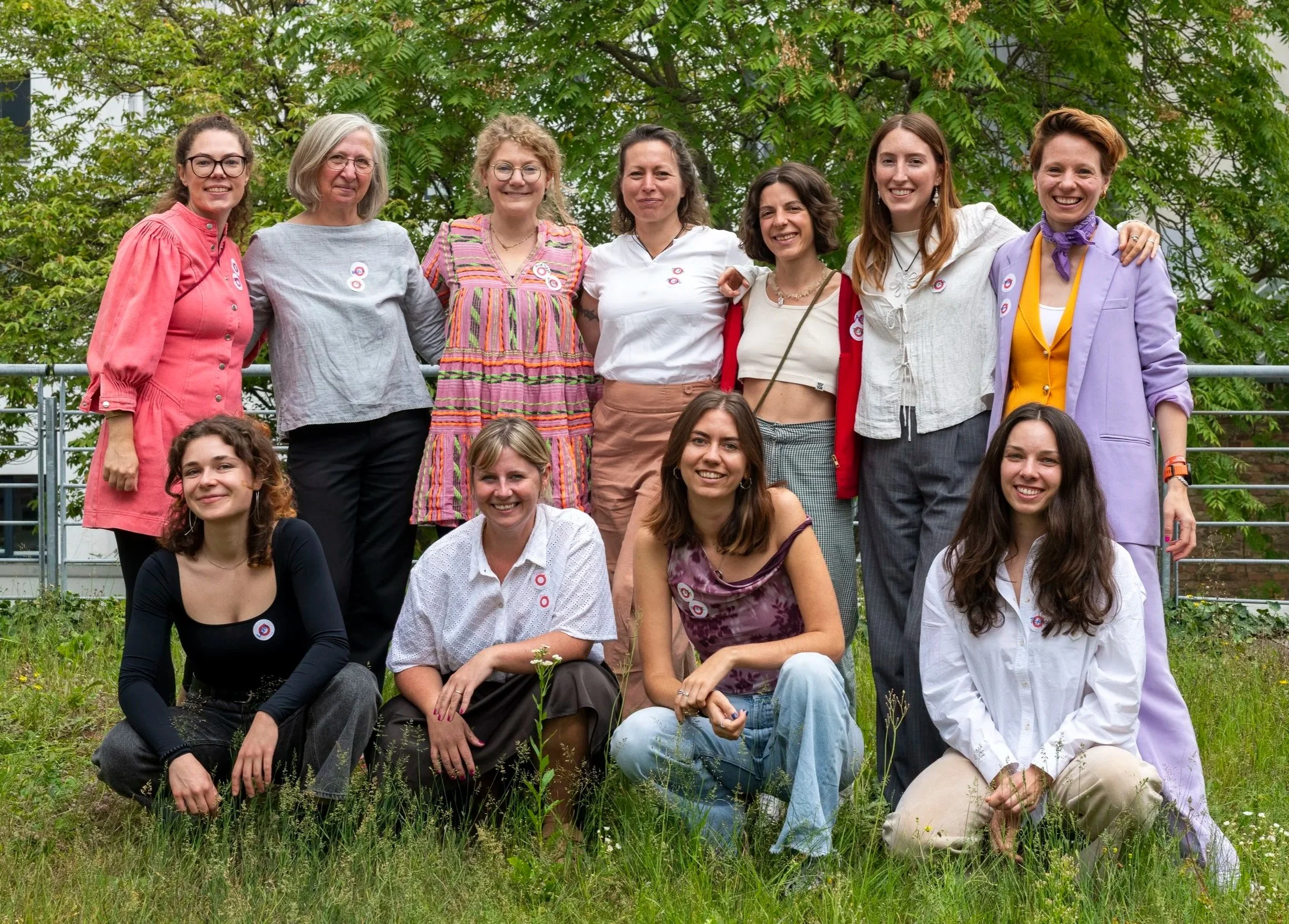 Gruppe von zwölf Frauen im Freien auf einer Wiese mit Bäumen im Hintergrund, einige stehen, einige sitzen, alle lächeln.