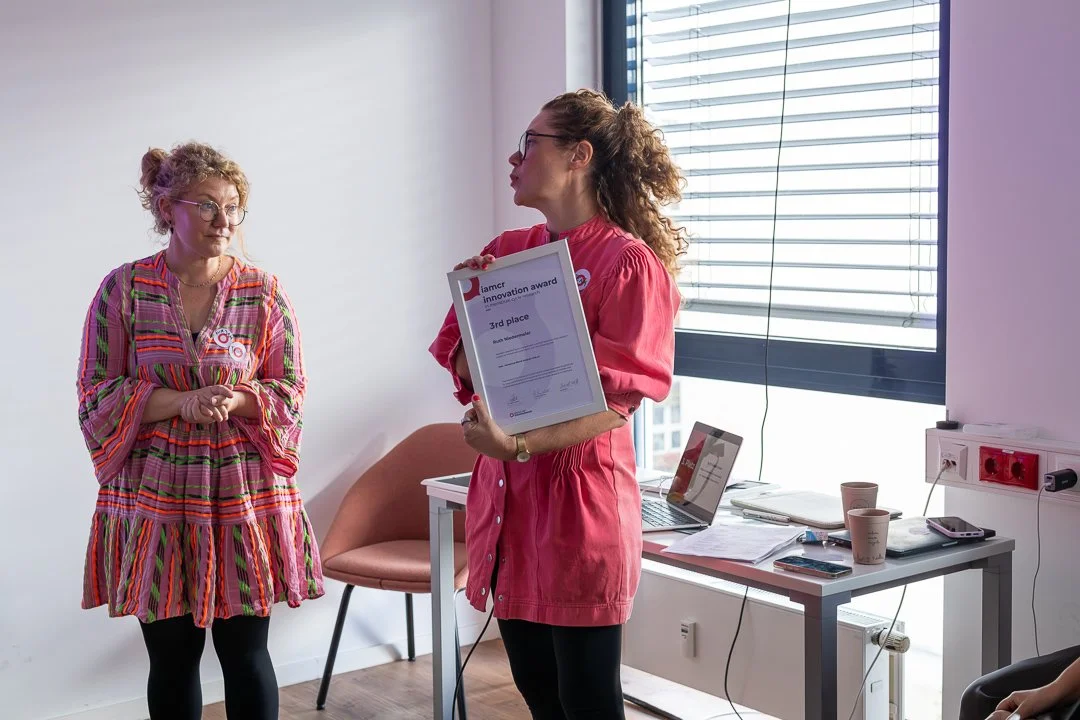 Zwei Frauen bei einer Preisübergabe in einem Büro, eine hält eine Urkunde für den 3. Platz beim Innovation Award. Es sind Schreibwaren, Kaffee und ein Laptop auf einem Tisch im Hintergrund.