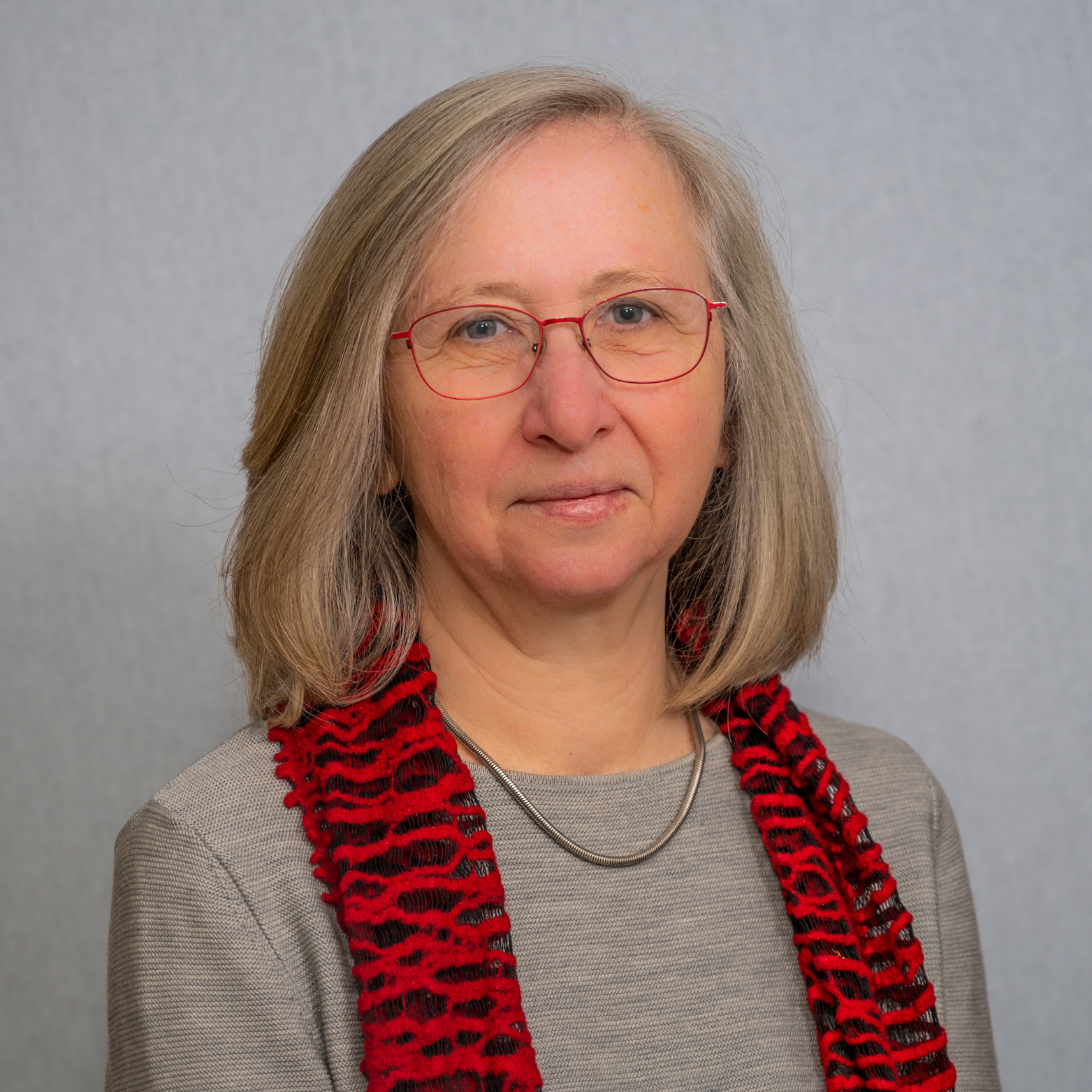 Porträt einer älteren Frau mit grauem Haar, rotem und schwarz gestreiftem Schal, grauem Pullover und roten Brillengestellen vor neutralem grauem Hintergrund.