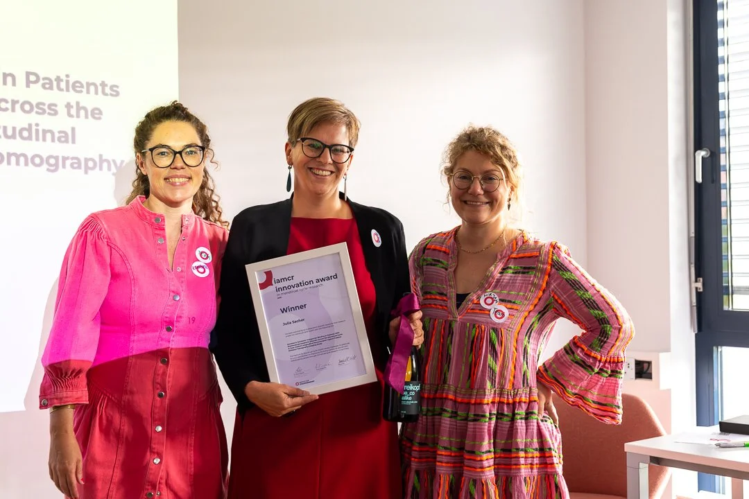 Drei Frauen stehen zusammen in einem Raum, eine von ihnen hält ein Zertifikat, die anderen beiden haben Buttons auf ihrer Kleidung. Die Frauen lächeln für die Kamera.