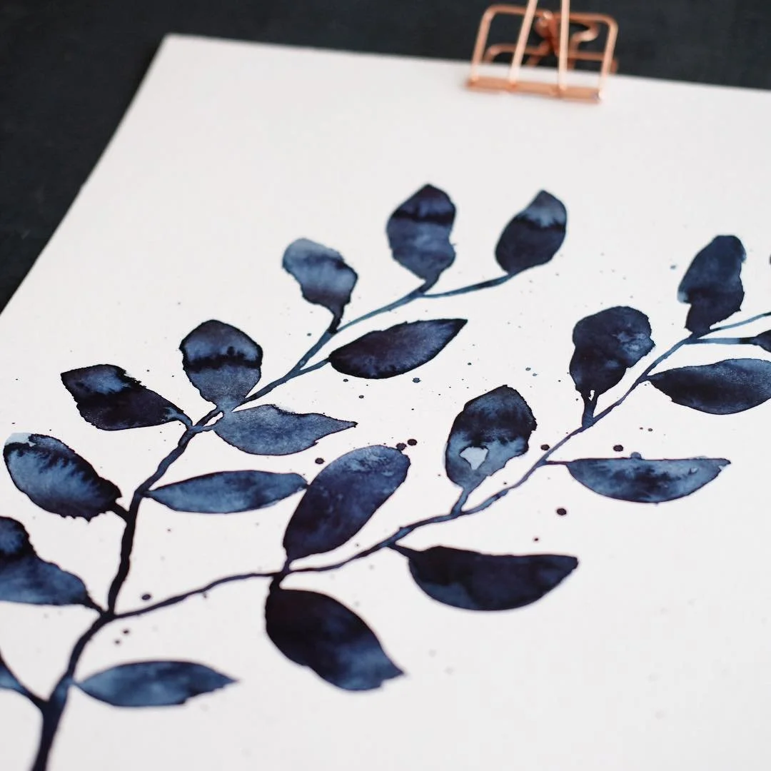 A watercolor painting of dark blue leaves on white paper held by a copper-colored clip.