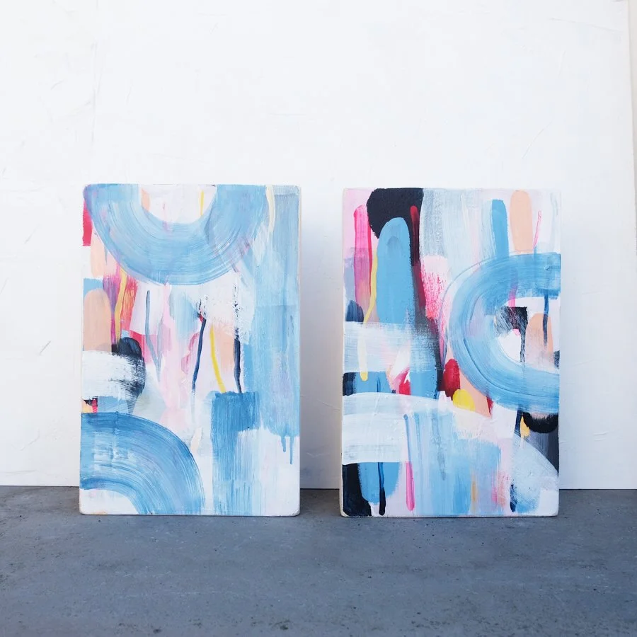 Zwei abstrakte Gemälde mit bunten, horizontal und vertikal verlaufenden Pinselstrichen in Blau, Pink, Schwarz, Weiß und Gelb, auf Holz-Leinwänden, auf einem Boden vor weißer Wand.